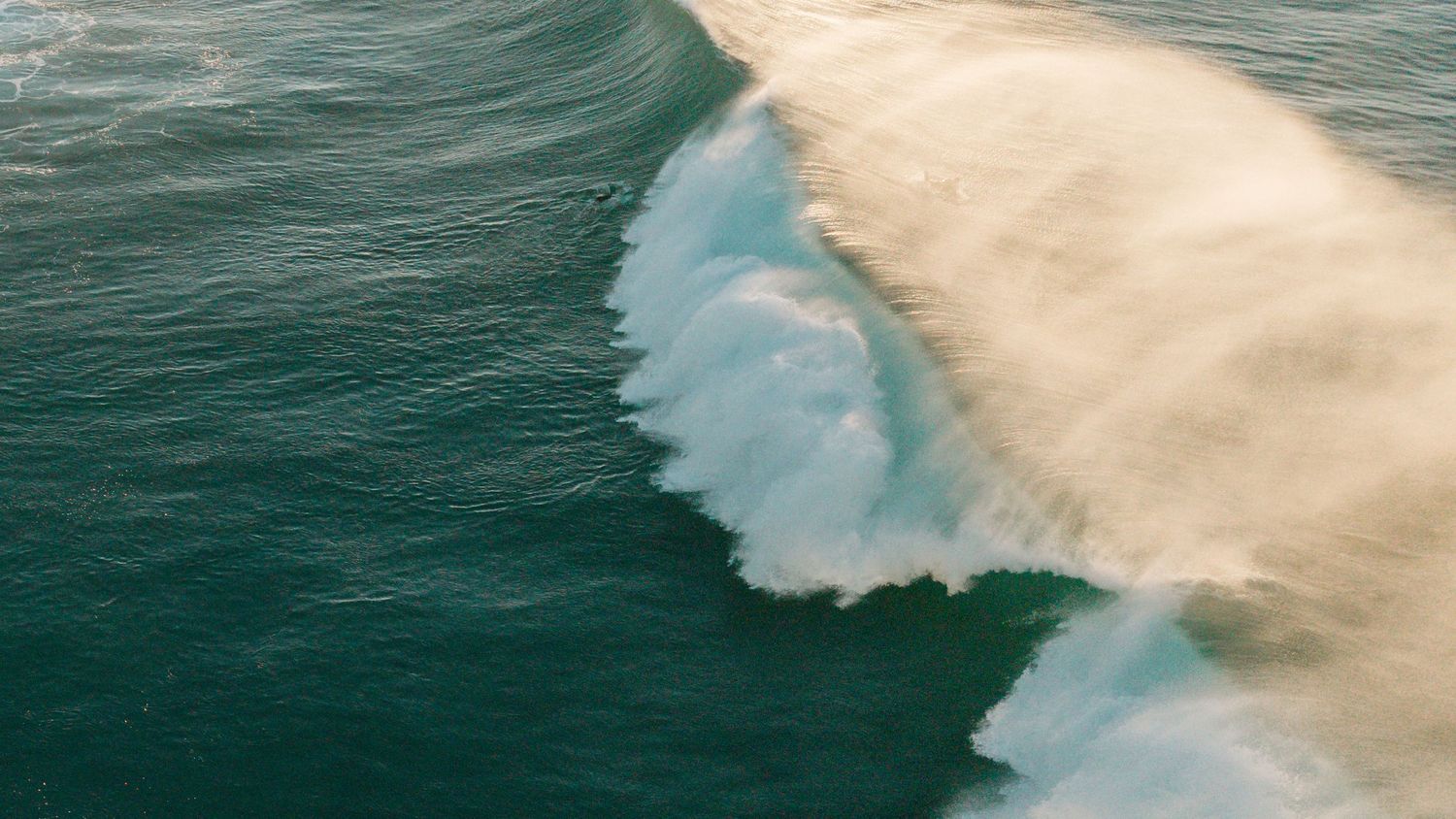 Aerial view of a massive ocean wave breaking with white foam and sandy brown water meeting deep teal sea.