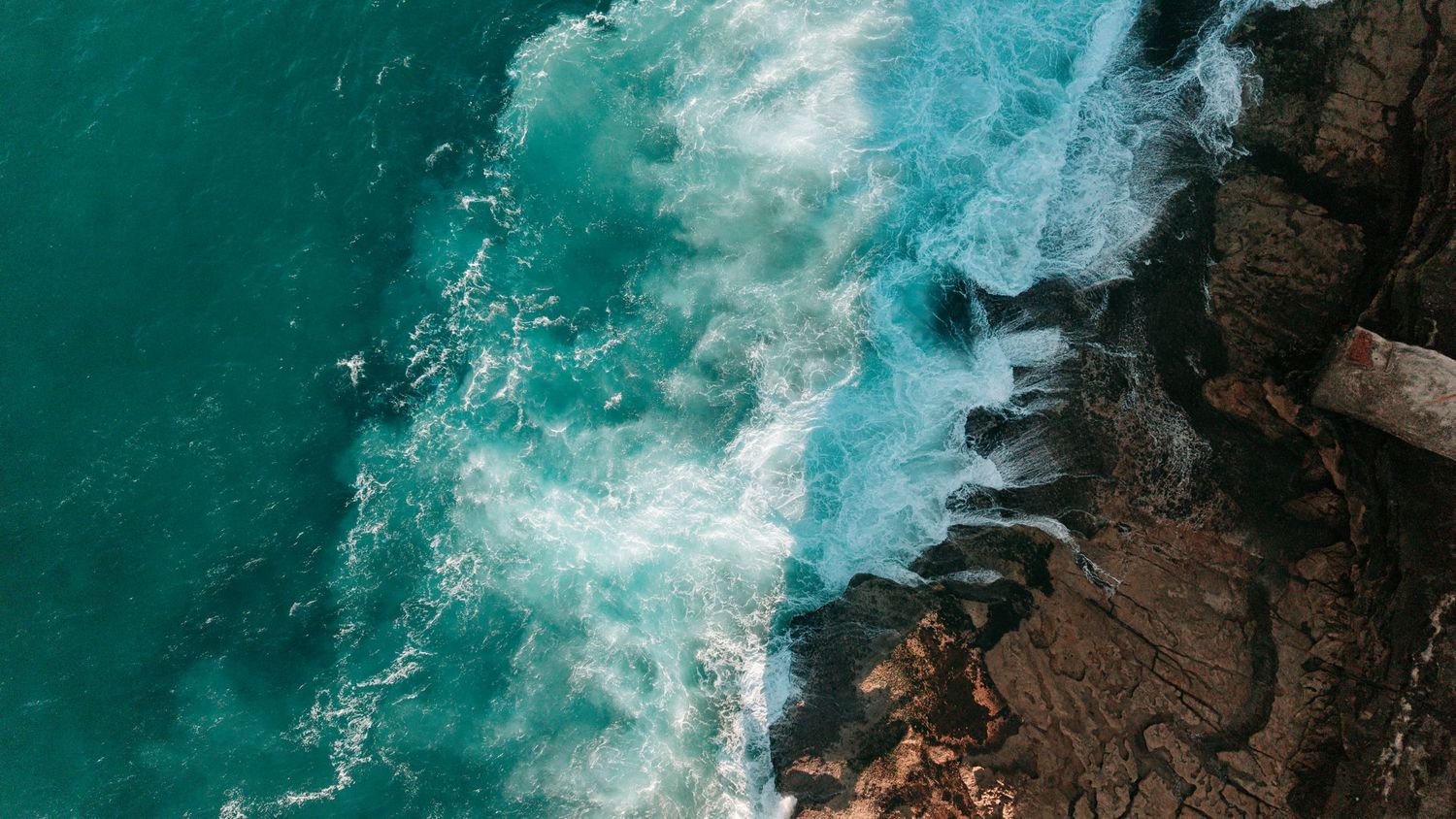 Aerial view of turquoise ocean waves crashing against rocky coastal cliffs with white sea foam.