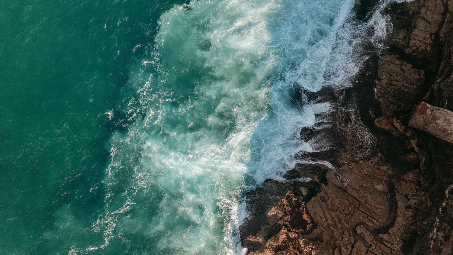Aerial view of turquoise ocean waves crashing against rugged rocky coastline, creating white sea foam.