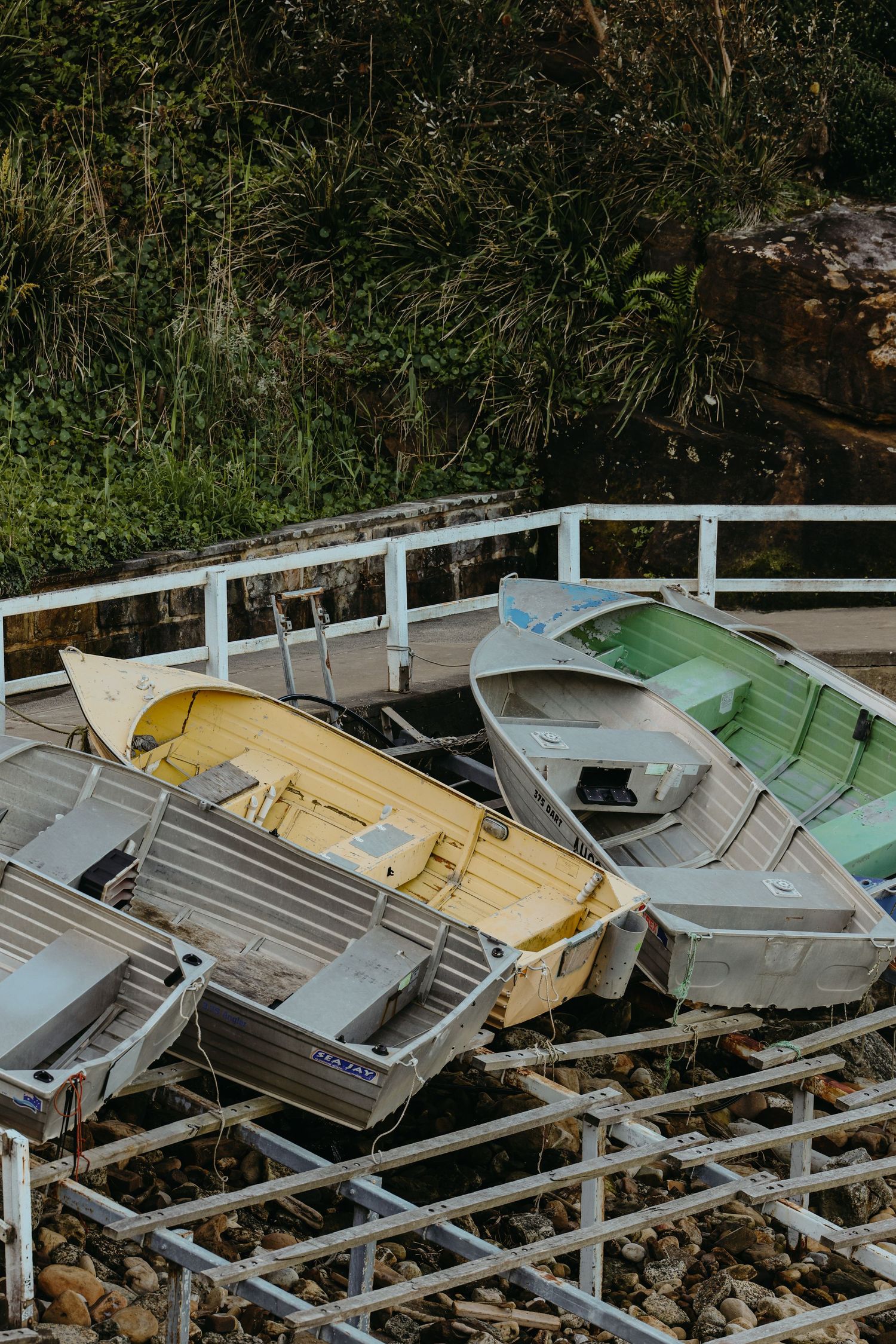 Row of weathered aluminum boats stacked on wooden racks near vegetation.