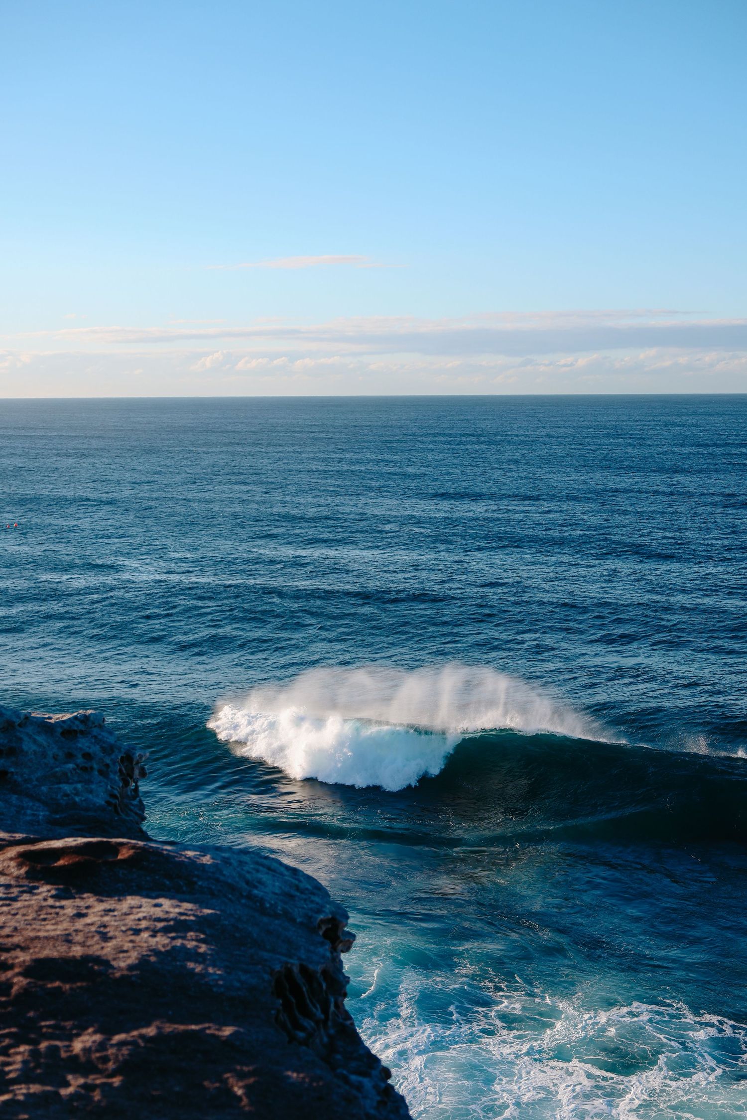 A powerful ocean wave crashes near rocky cliffs under a clear blue sky with calm open sea in the background.