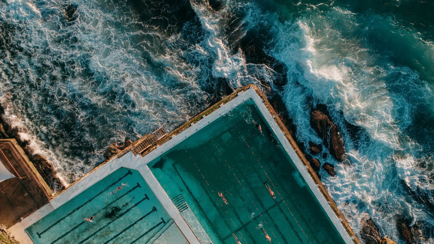 Aerial view of Bondi Icebergs ocean pool with turquoise water beside crashing waves on rocky coastline.