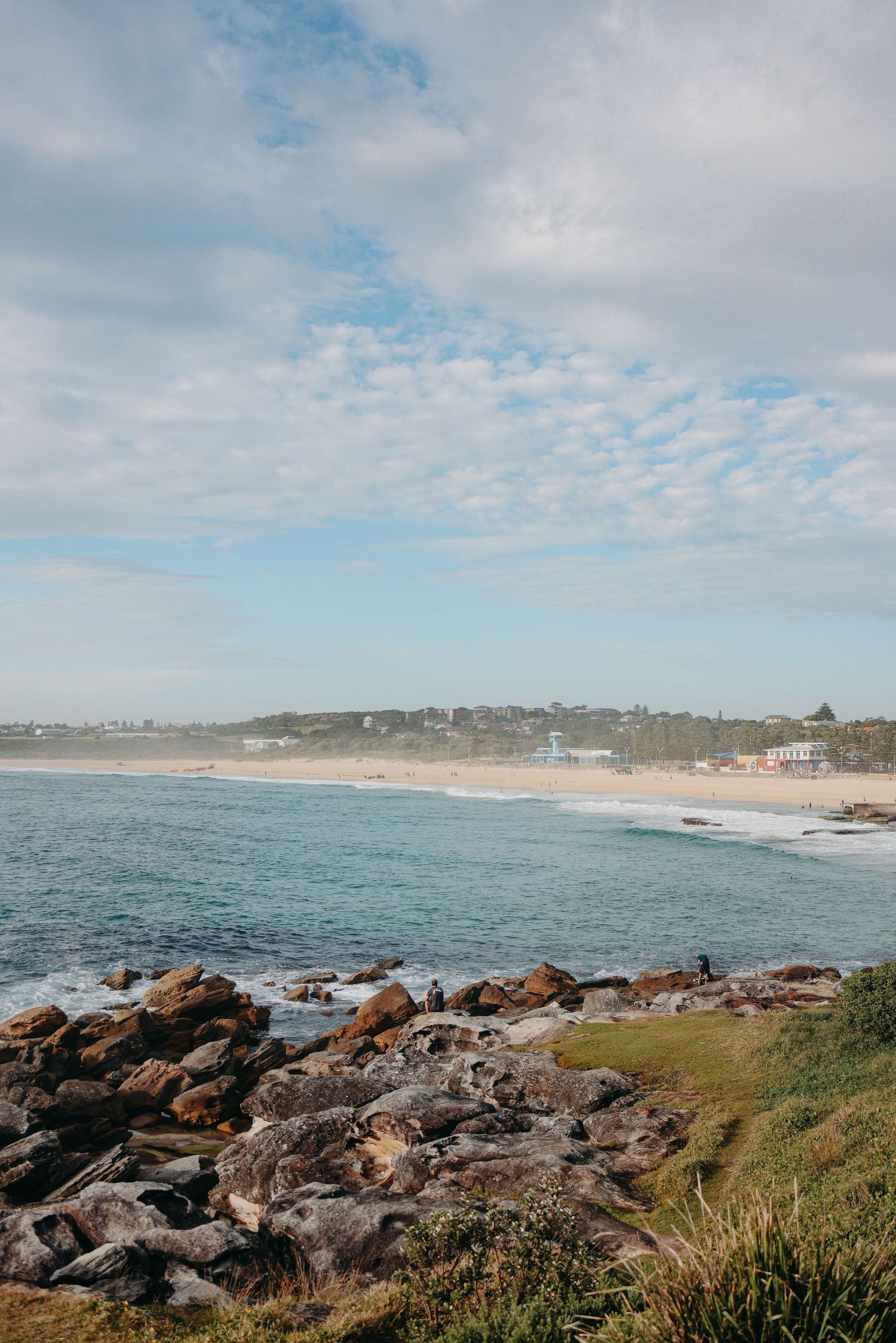 Rocky coastal viewpoint overlooking blue ocean with beach town visible in the distance.