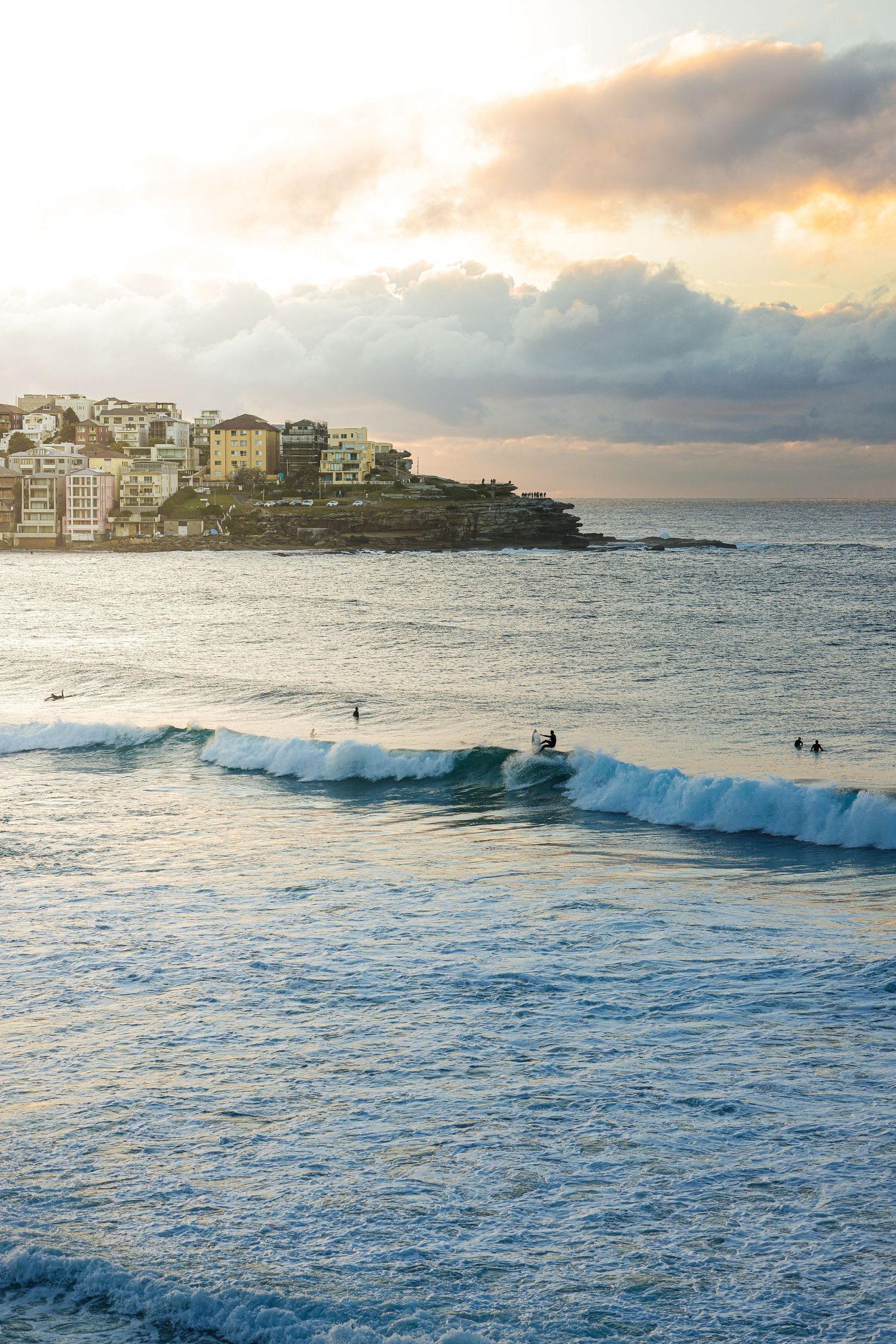 Coastal cityscape with crashing waves and waterfront buildings during golden hour.