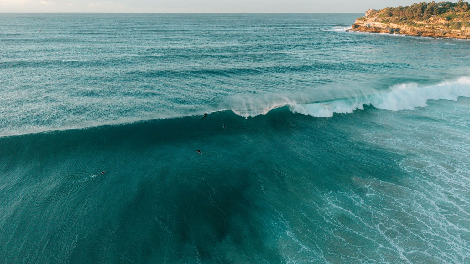 Aerial view of a large turquoise ocean wave breaking near a rocky coastline under a clear blue sky.