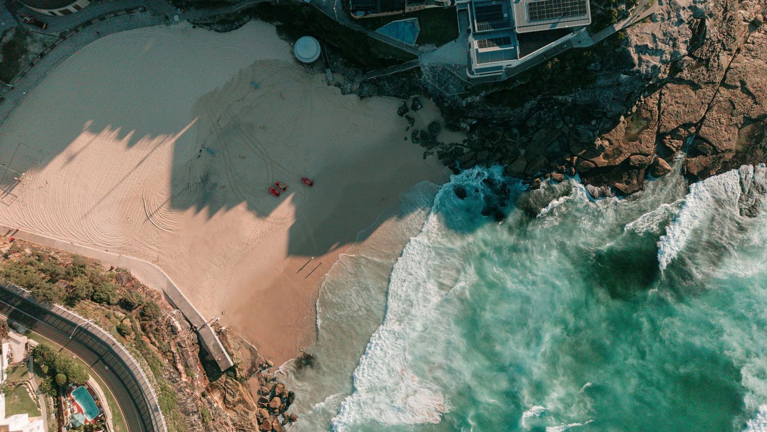 Aerial view of a sandy beach meeting turquoise ocean waves beside rocky cliffs and coastal buildings.