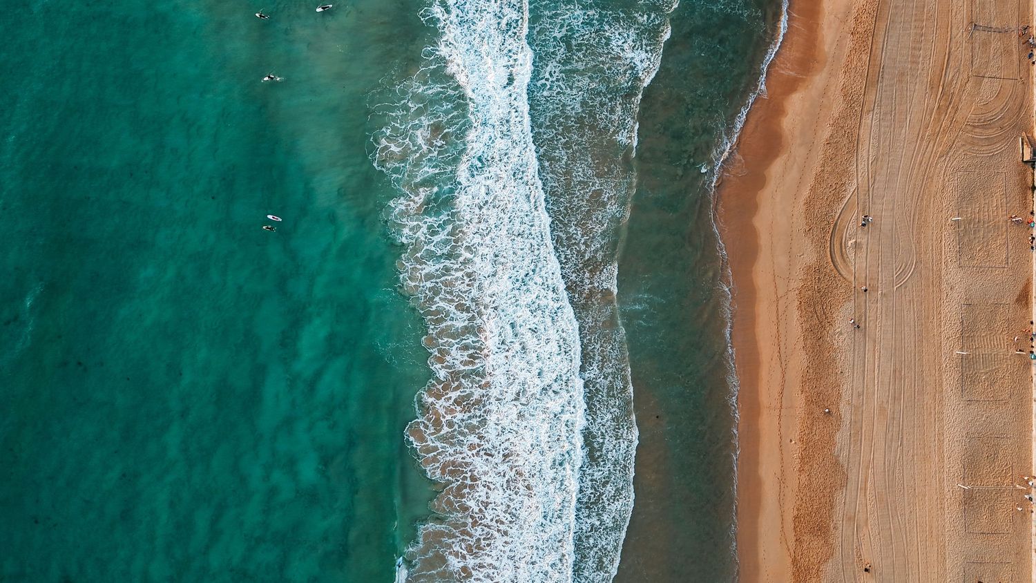 Drone shot of white surf meeting sandy beach with turquoise ocean waters.
