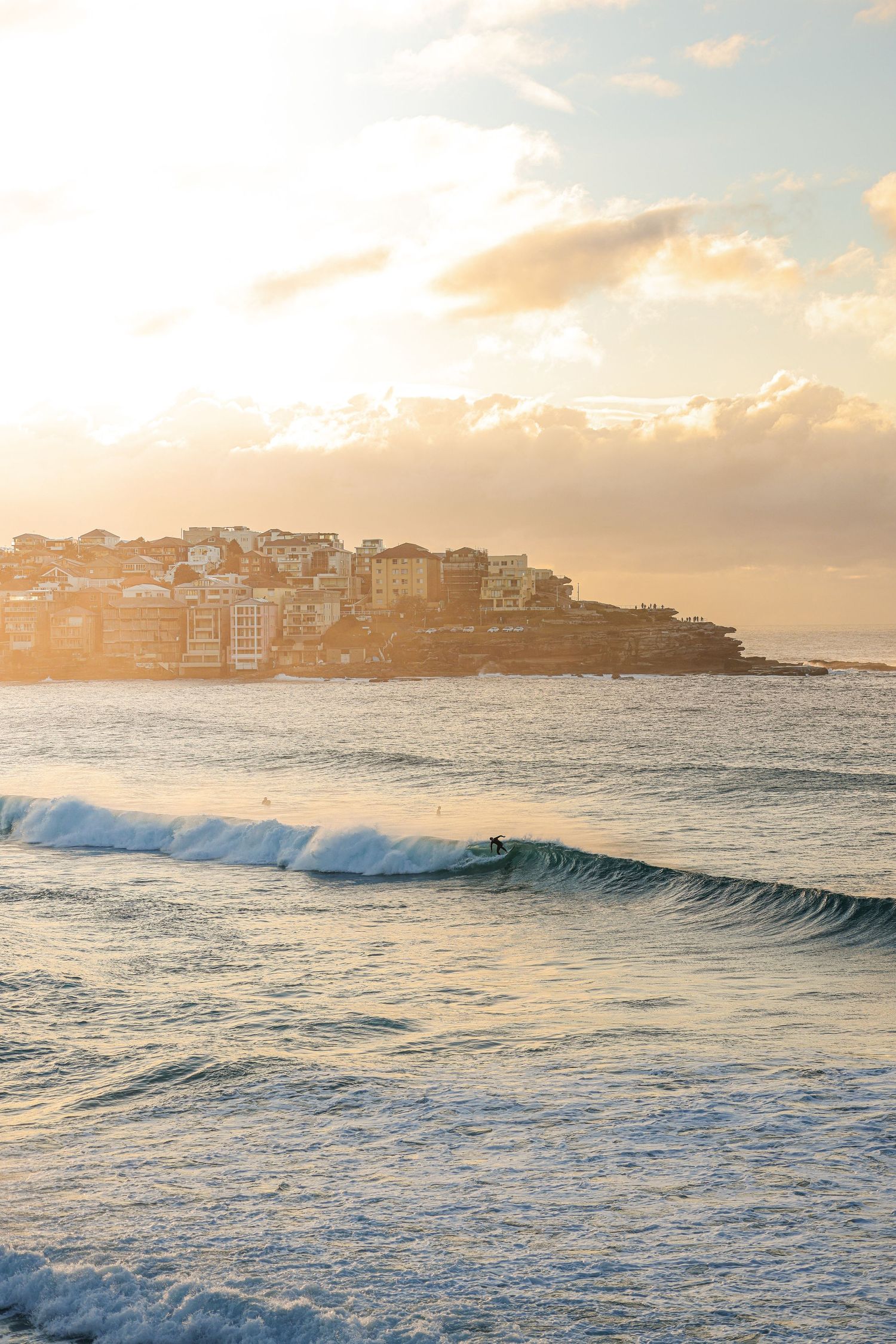 Coastal cityscape at sunset with breaking ocean waves and buildings silhouetted against a golden sky.