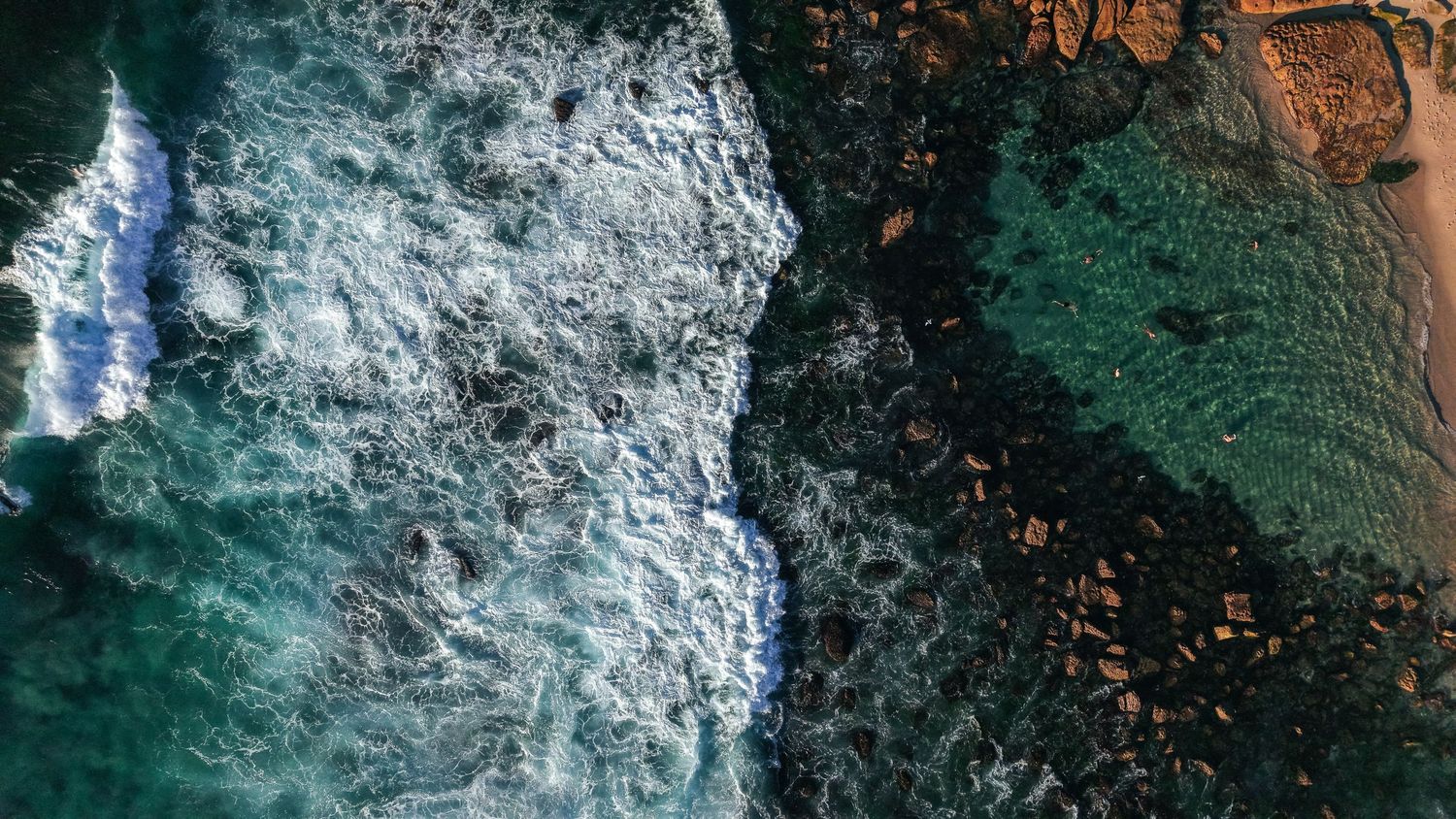 Aerial view of ocean waves crashing against rocky coastline with turquoise water and white sea foam.