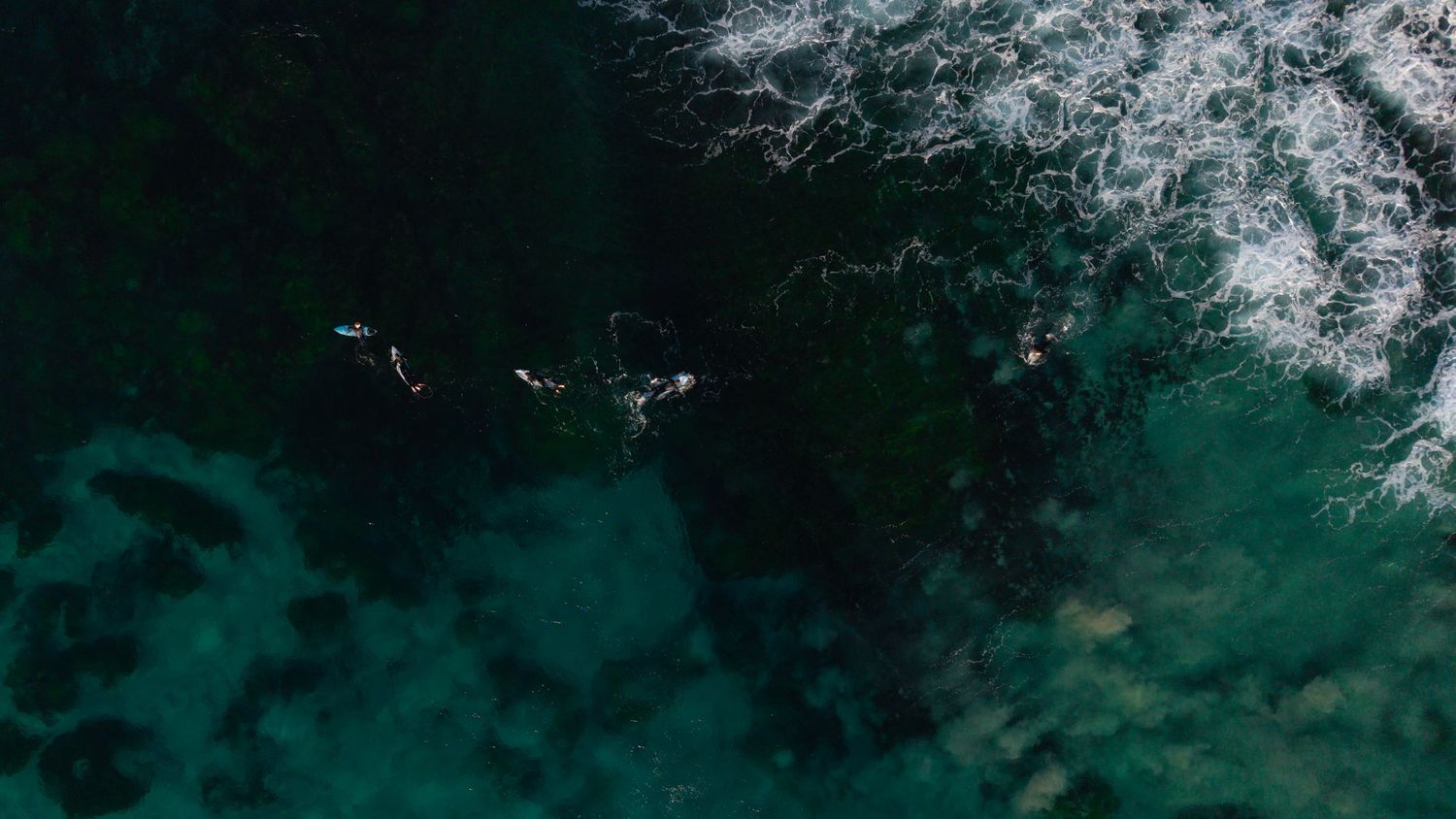 Aerial drone view of surfers riding waves in deep turquoise ocean water with rocky reef visible below.