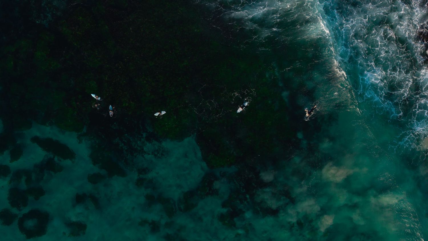 Aerial view of surfers paddling on clear turquoise ocean water near rocky reef formations.