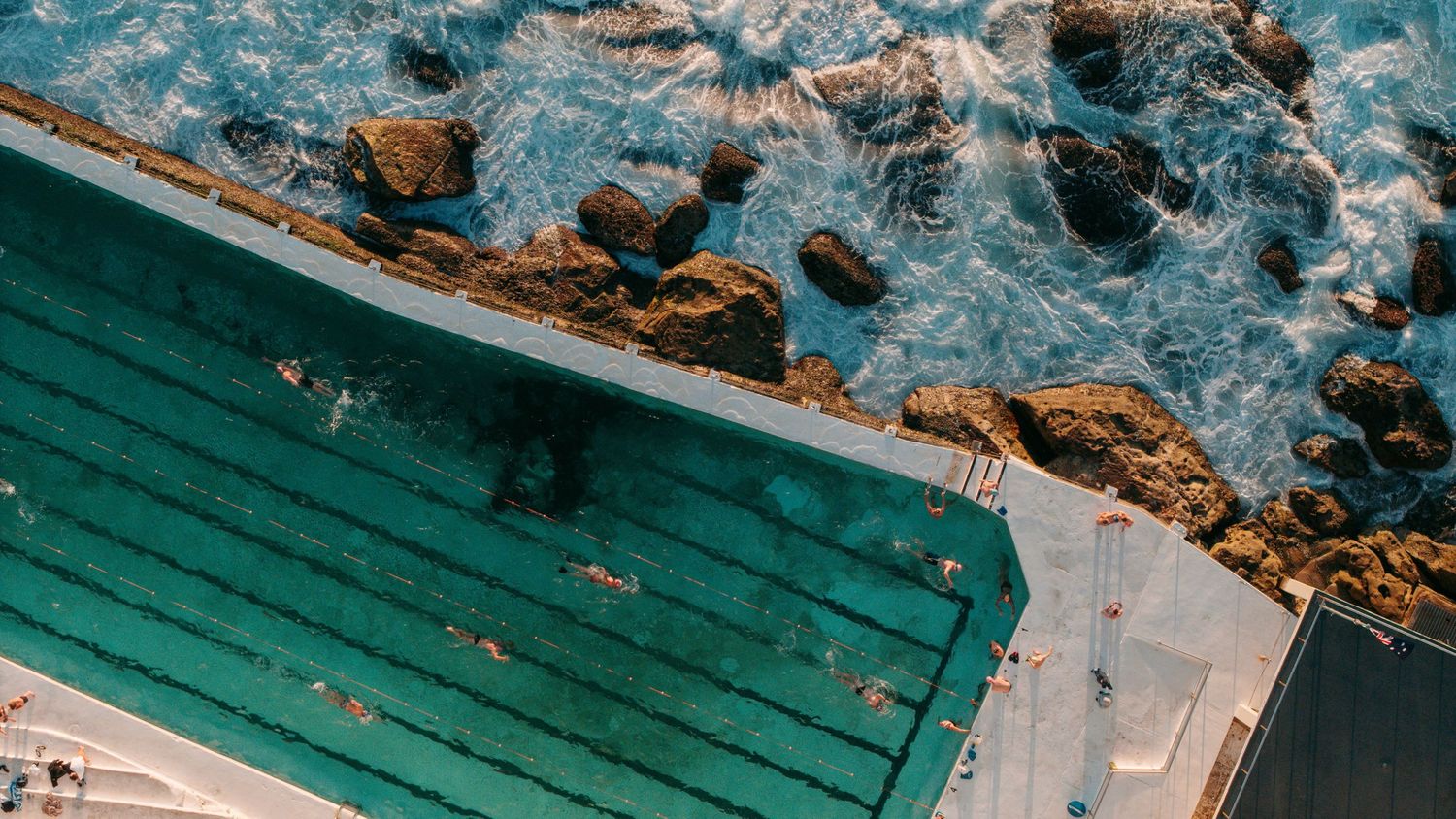 Aerial view of Bondi Icebergs ocean pool with swimmers, surrounded by rocky coastline and turquoise waves.