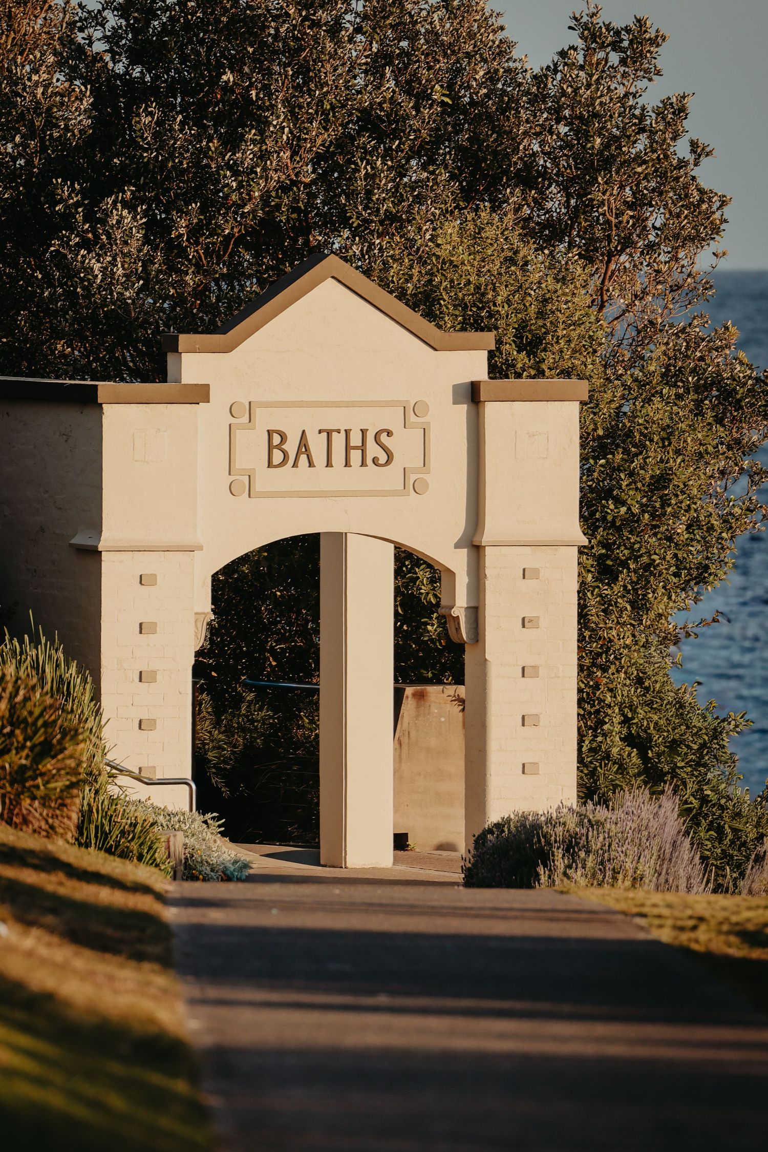 White stone archway entrance marked 'BATHS' along a waterfront path.