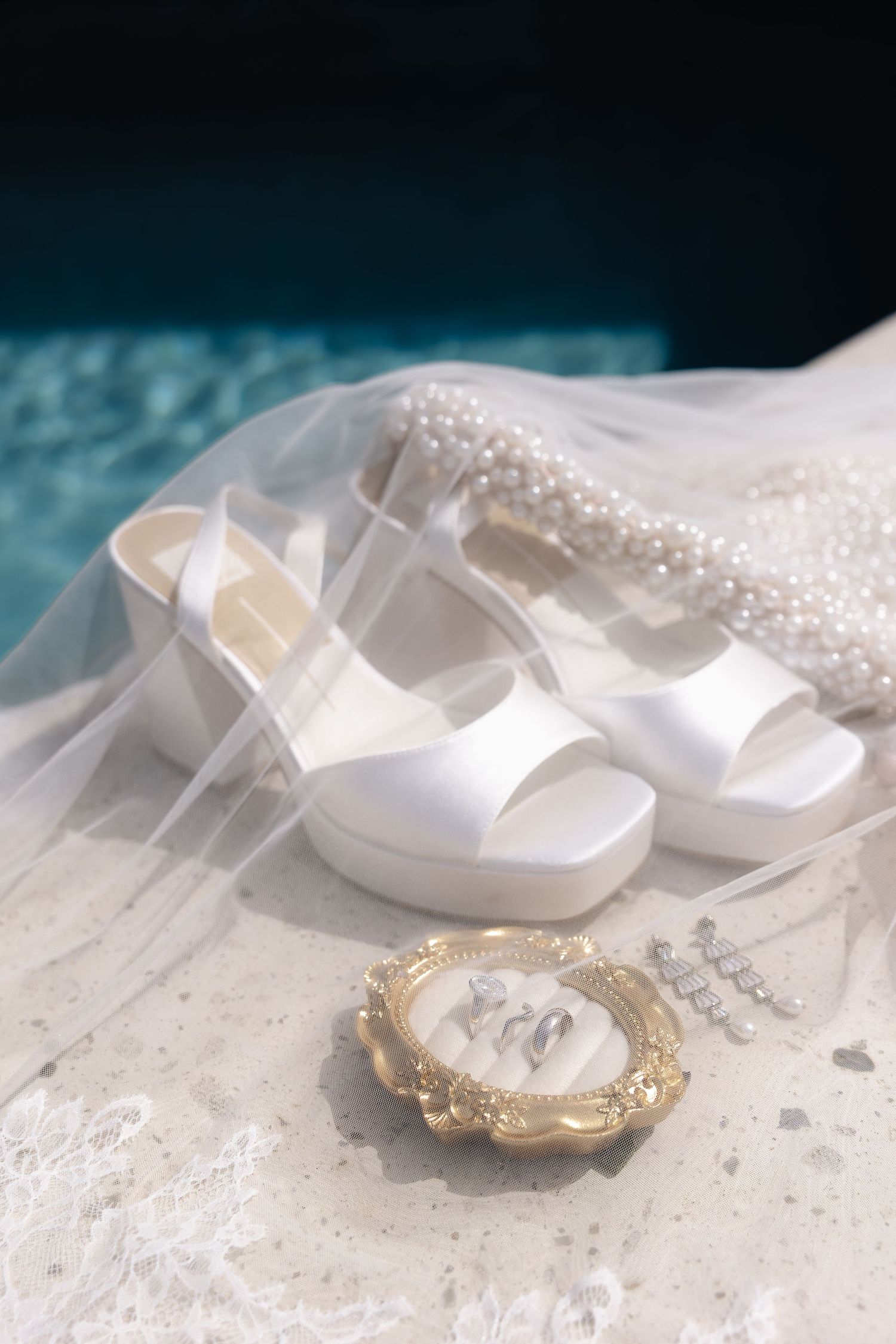 White platform bridal heels with veil, pearl accessories, and gold jewelry dish on marble surface near pool.