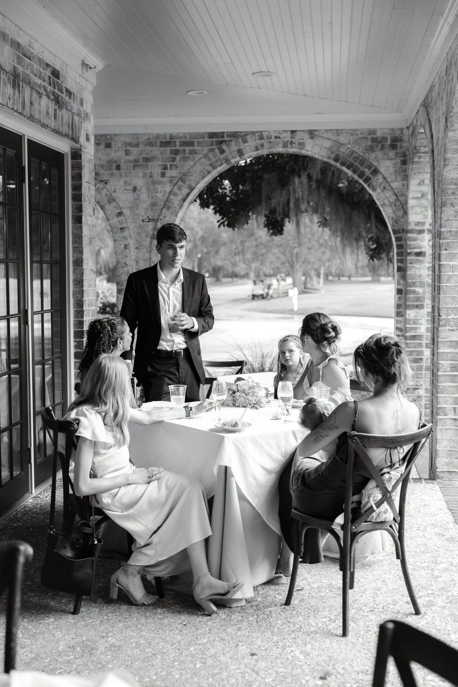 Black and white photo of wedding guests gathered at a table on a brick porch, with a man standing and raising a glass.