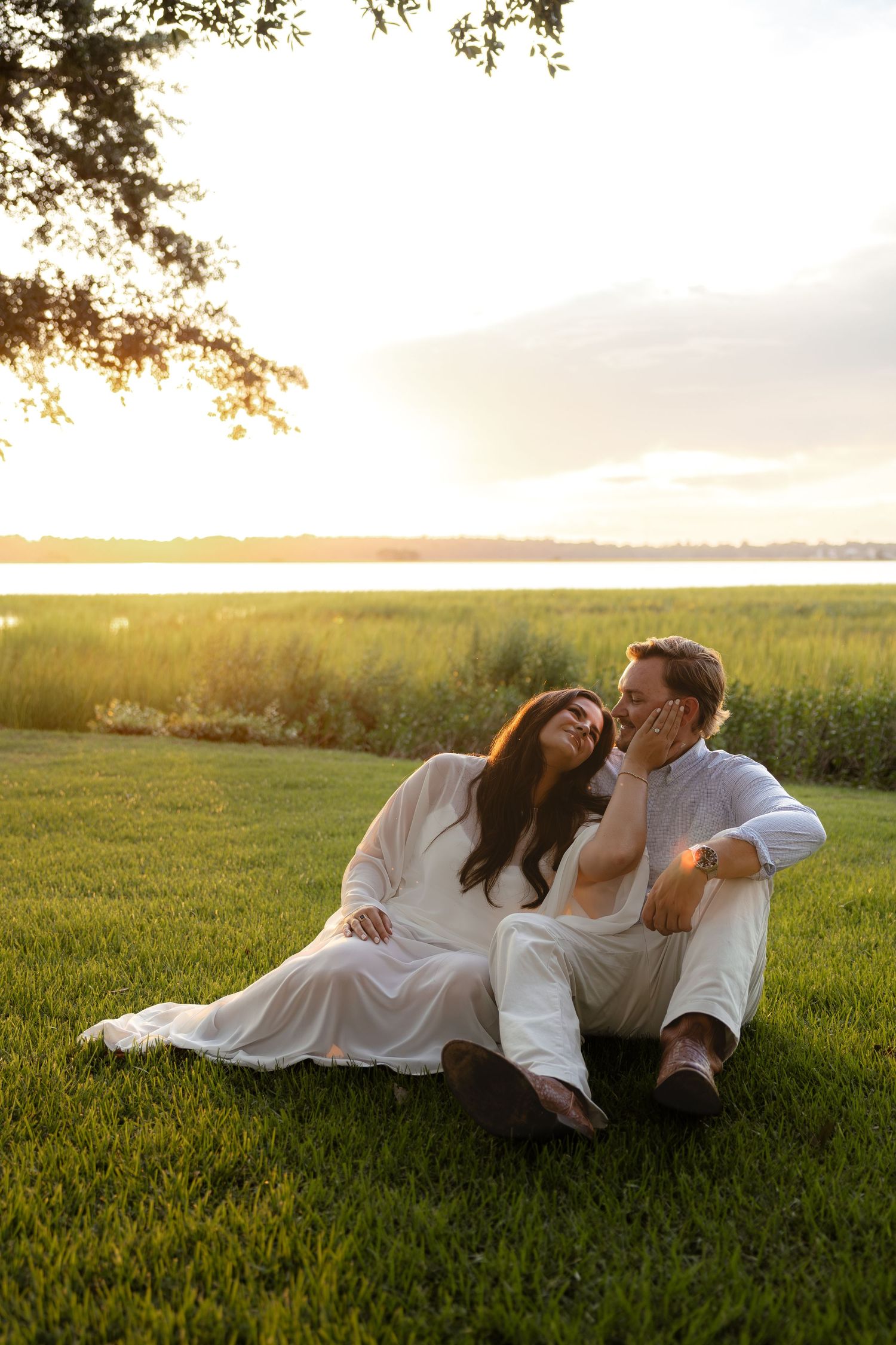 Couple in white outfits sitting on green grass by a marsh at golden hour sunset, sharing a romantic moment.