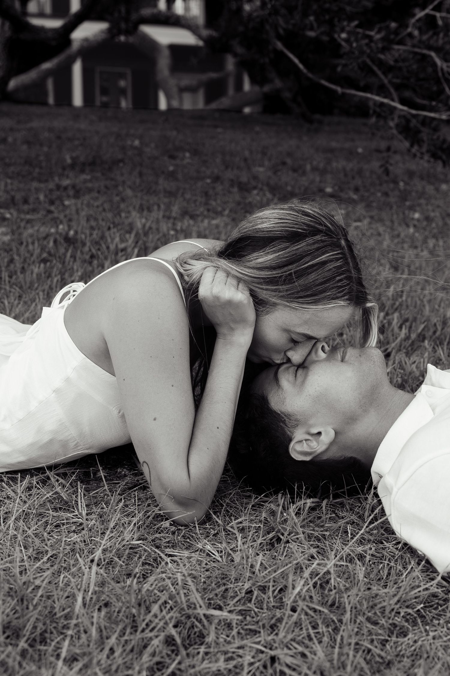 Black and white romantic photo of a couple lying on grass sharing a tender kiss during engagement shoot.