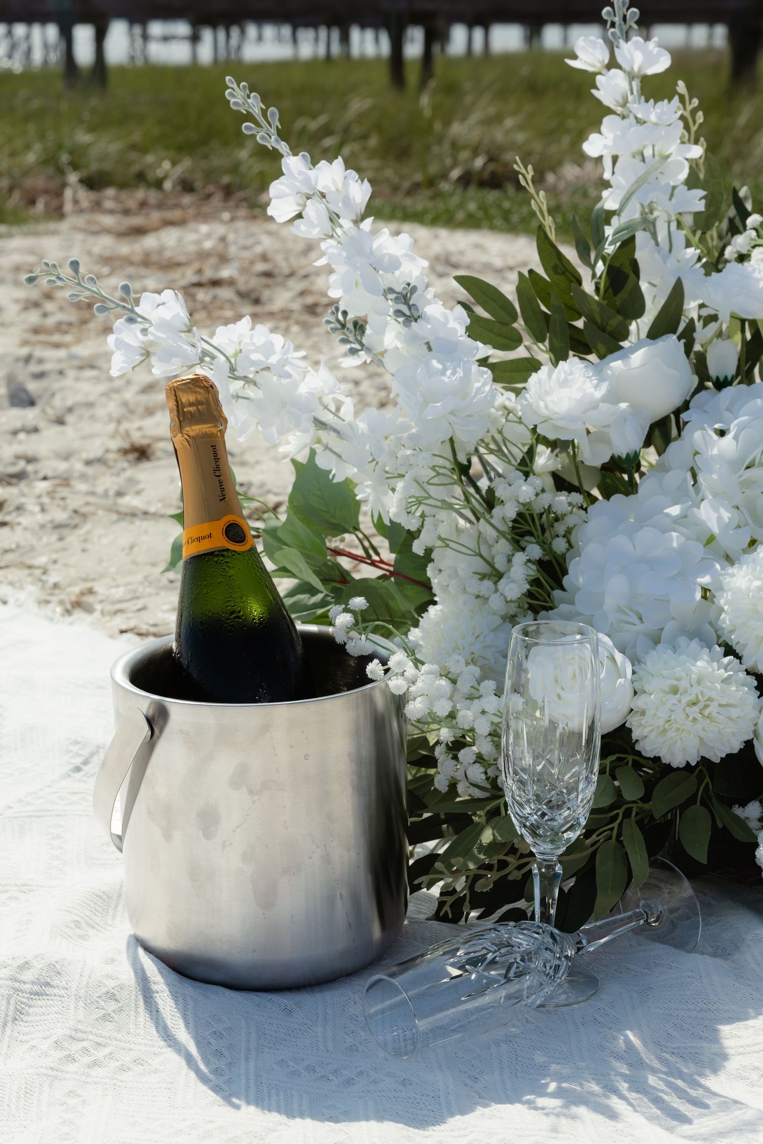 Champagne bottle in ice bucket with crystal flute and white floral arrangement outdoors.