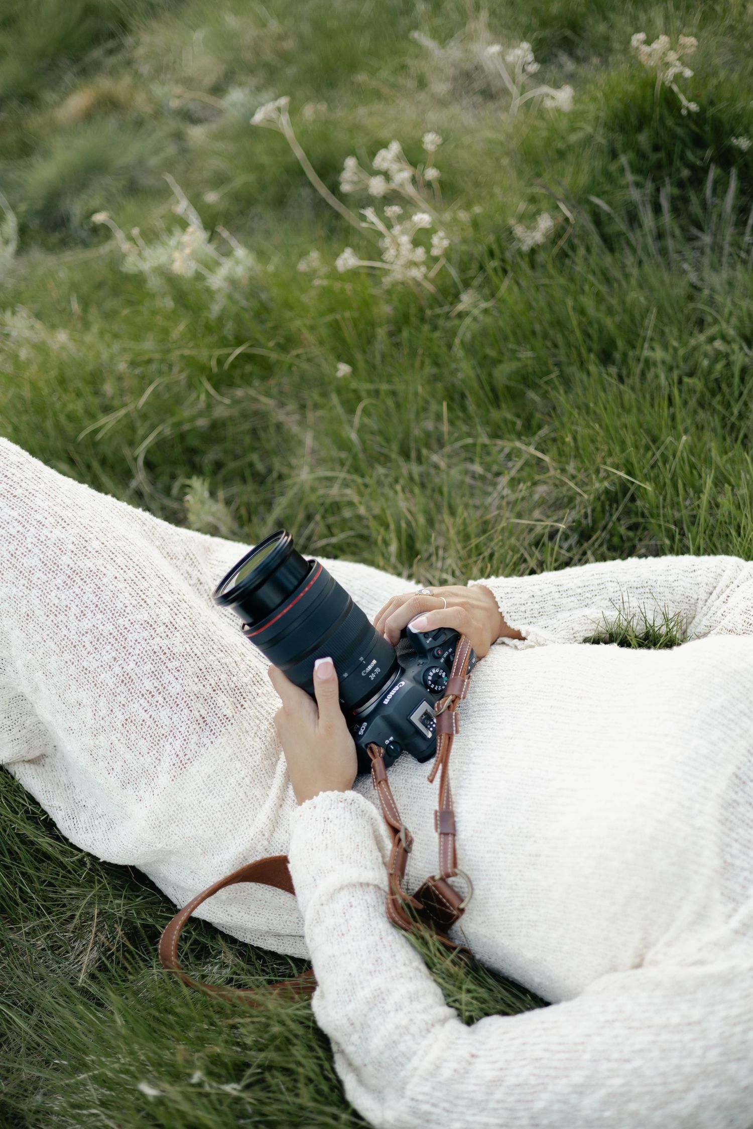 Person in white knit sweater lying in green grass holding DSLR camera with leather strap.