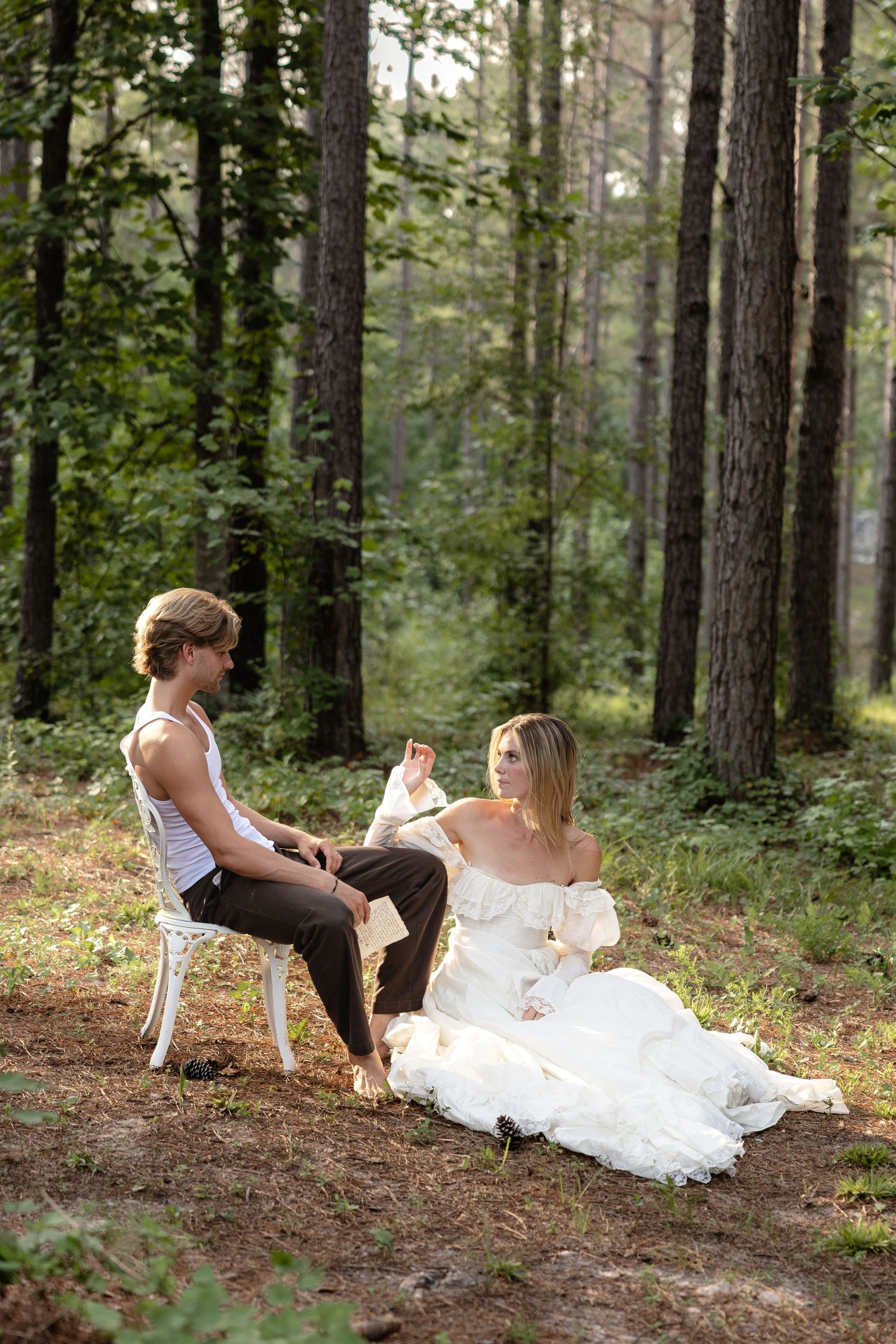 Bride in white gown sits on forest floor beside groom on chair, sharing a romantic moment among tall trees.