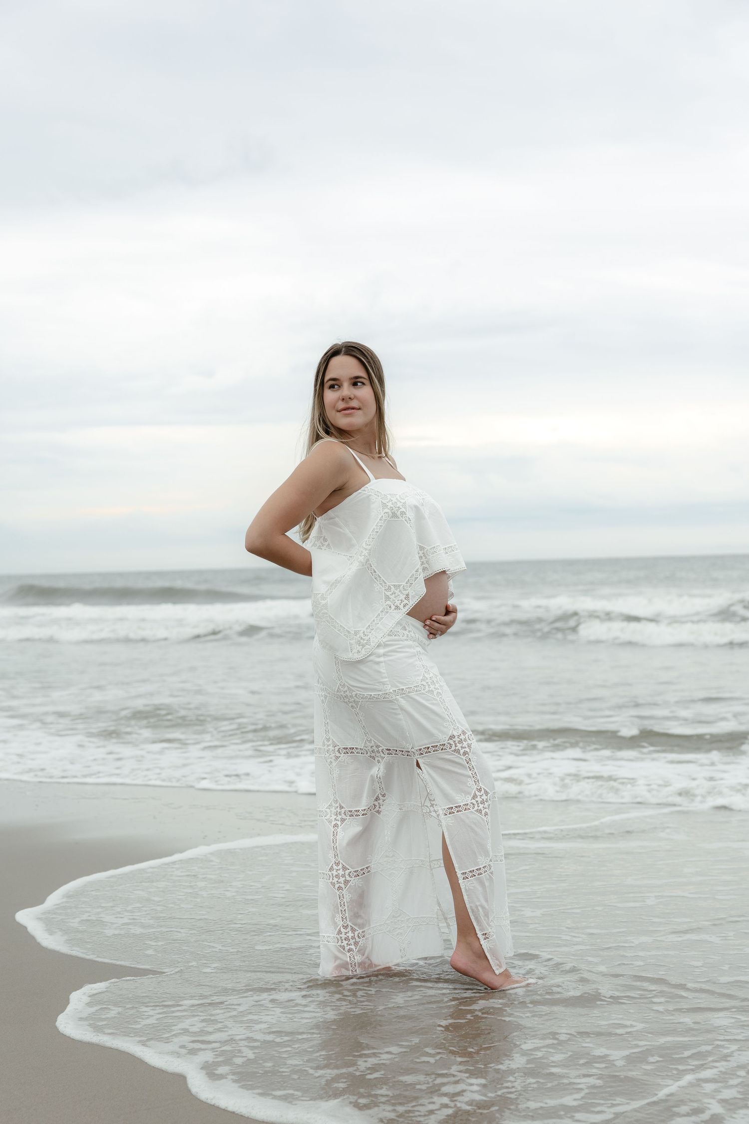 Pregnant woman in white lace dress standing in ocean waves on an overcast beach.