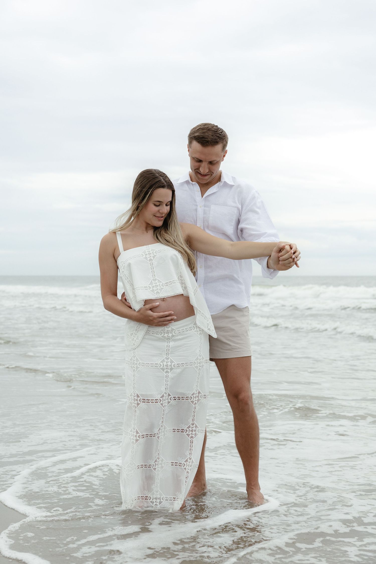 Pregnant couple standing in ocean waves, man holding woman's hand as she cradles her baby bump in white outfits.