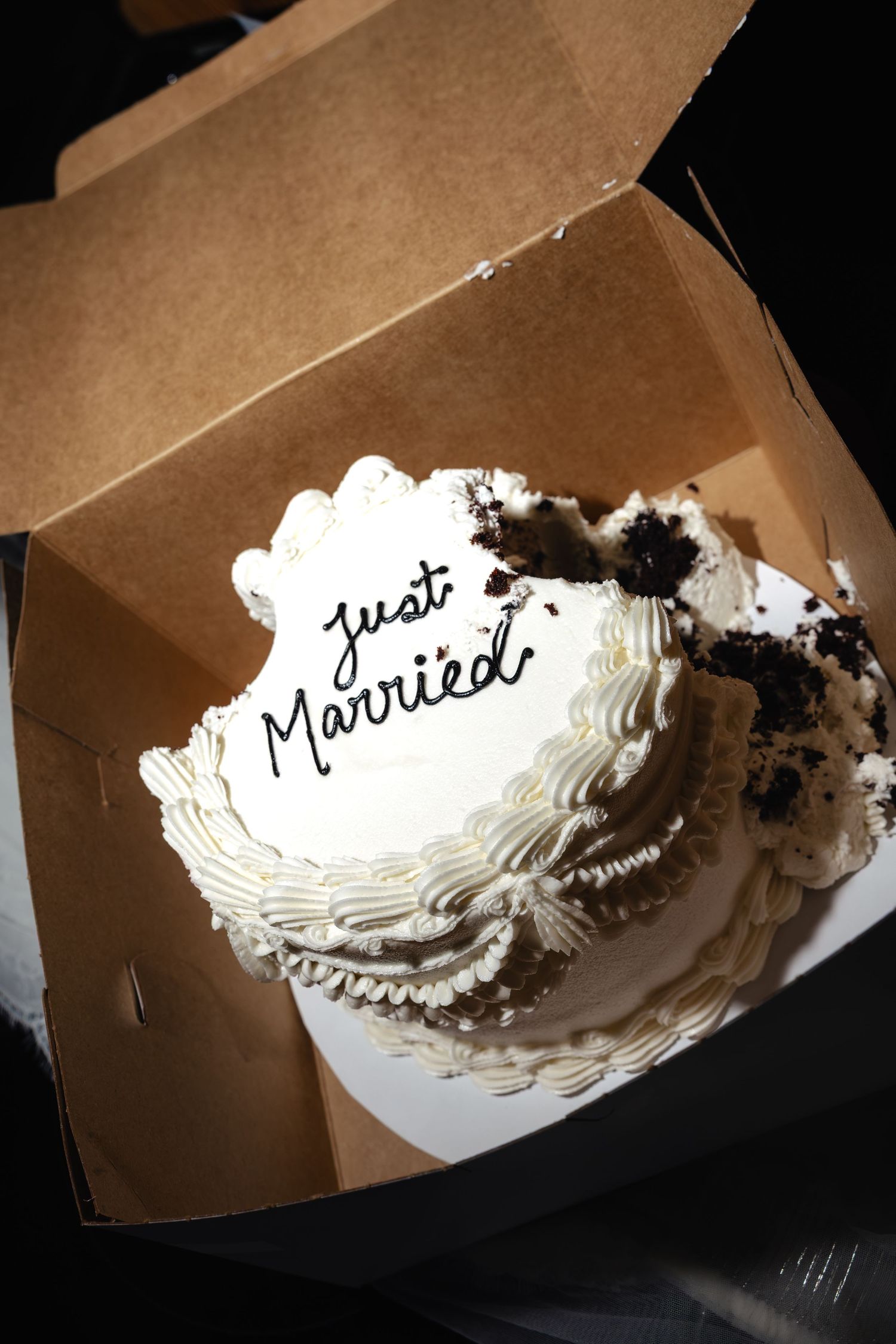 A white wedding cake with Just Married inscription in a cardboard box, with a slice removed revealing chocolate interior.