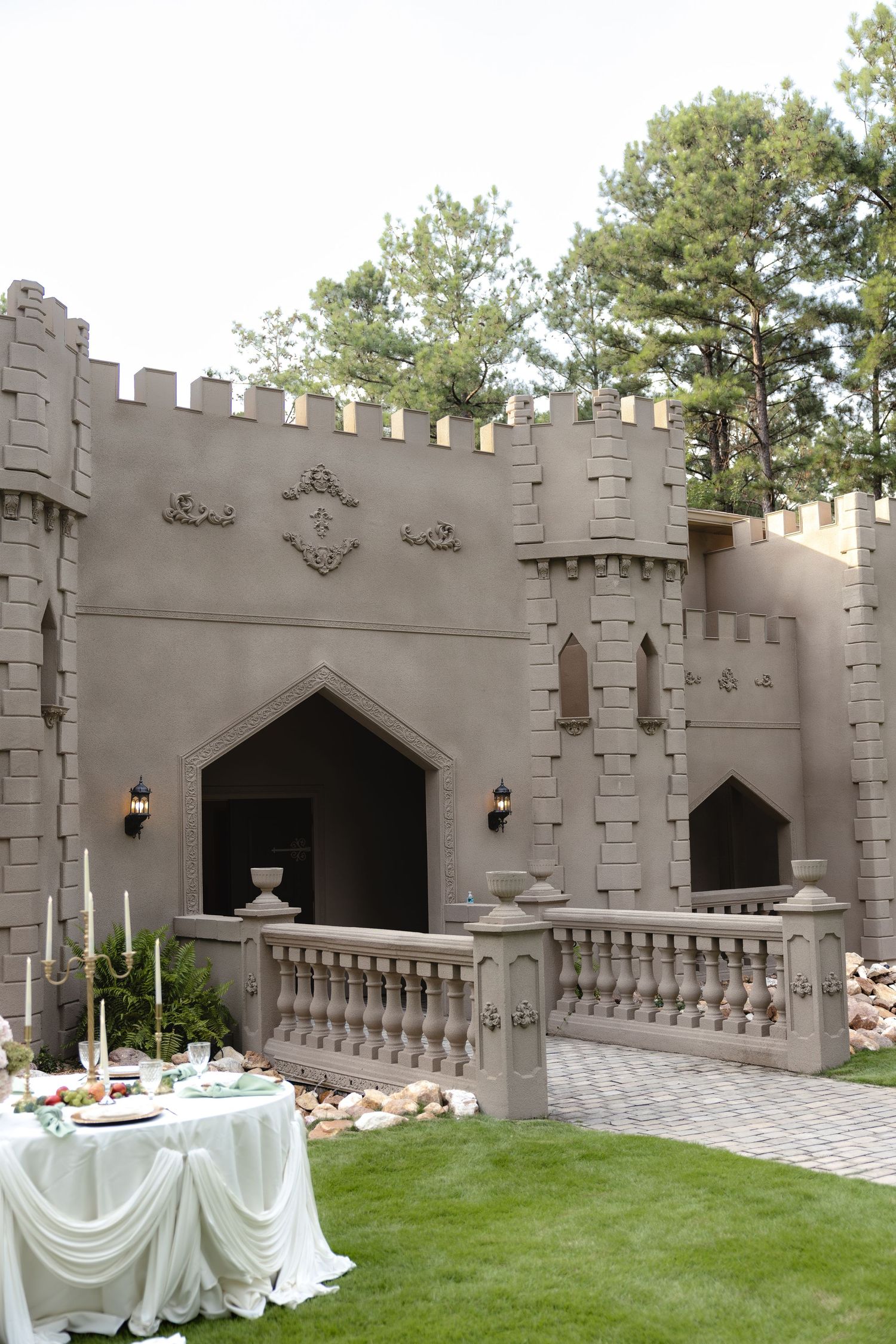 Castle-style wedding venue with battlements, ornate stone balustrade, candlelit tables, and lush green lawn surrounded by tall pine trees.