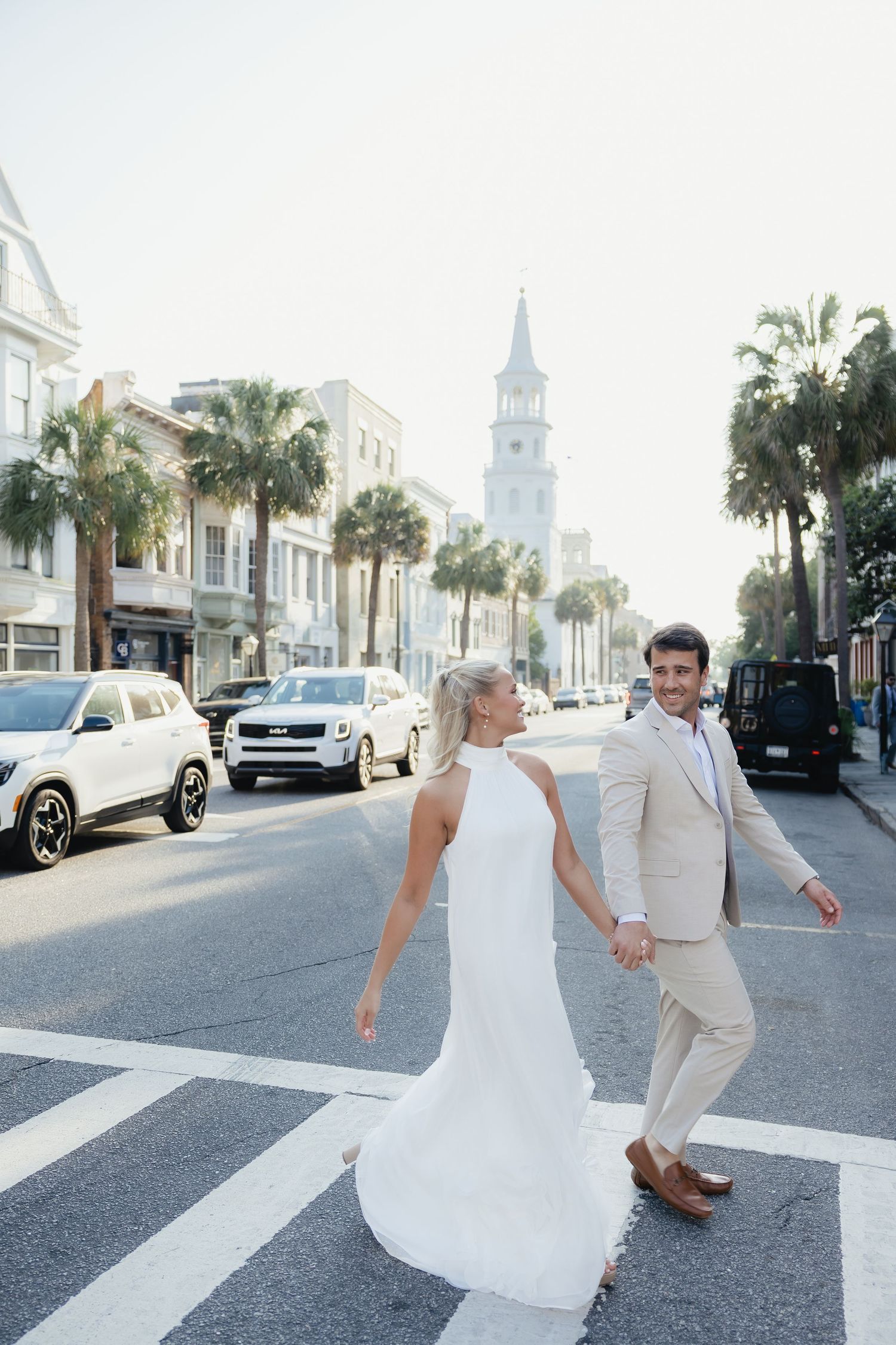 Bride and groom crossing a sunny Charleston street holding hands with palm trees and church steeple behind them.