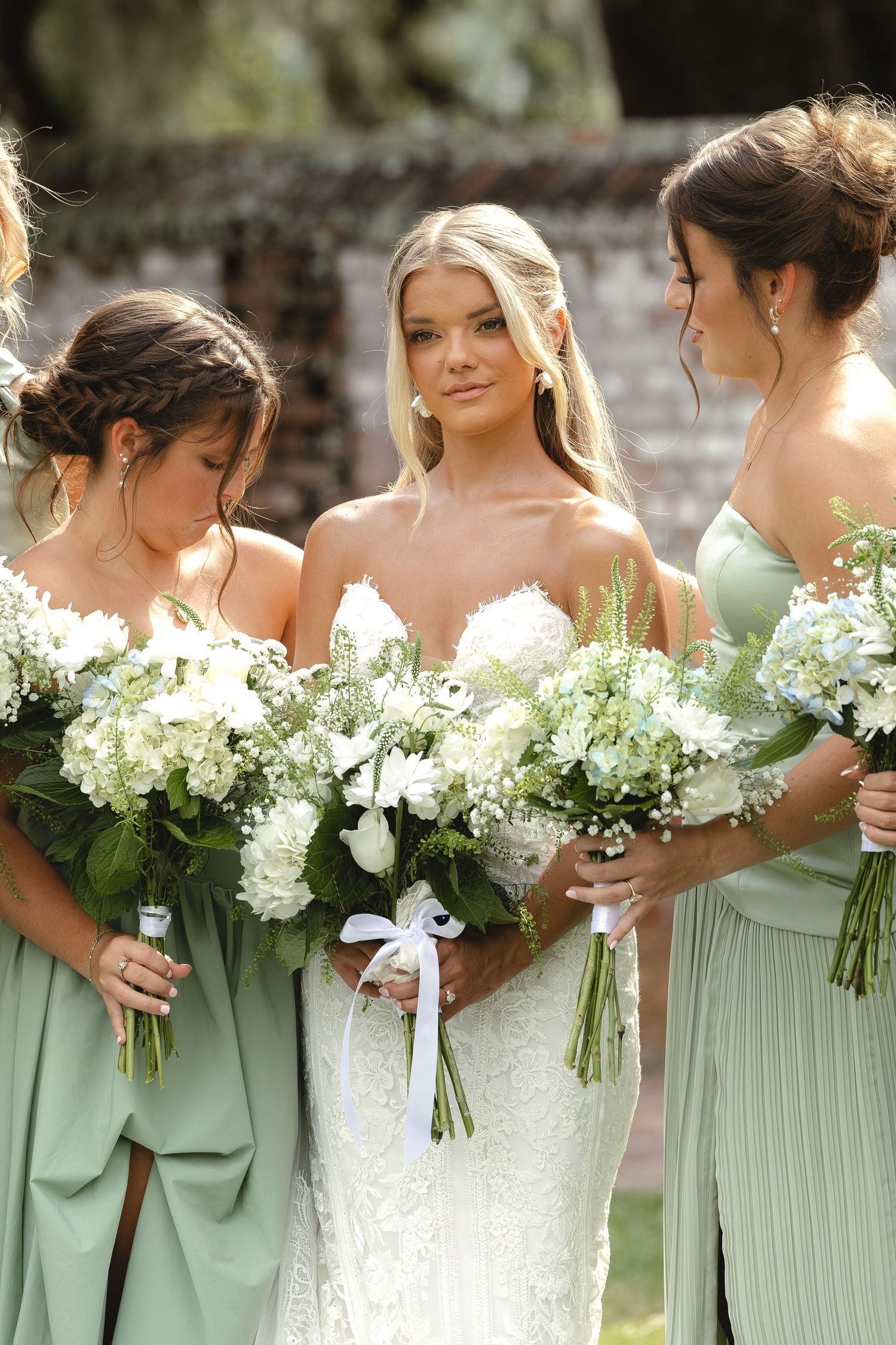 Bride in white lace gown with bridesmaids in sage green dresses holding white floral bouquets outdoors.