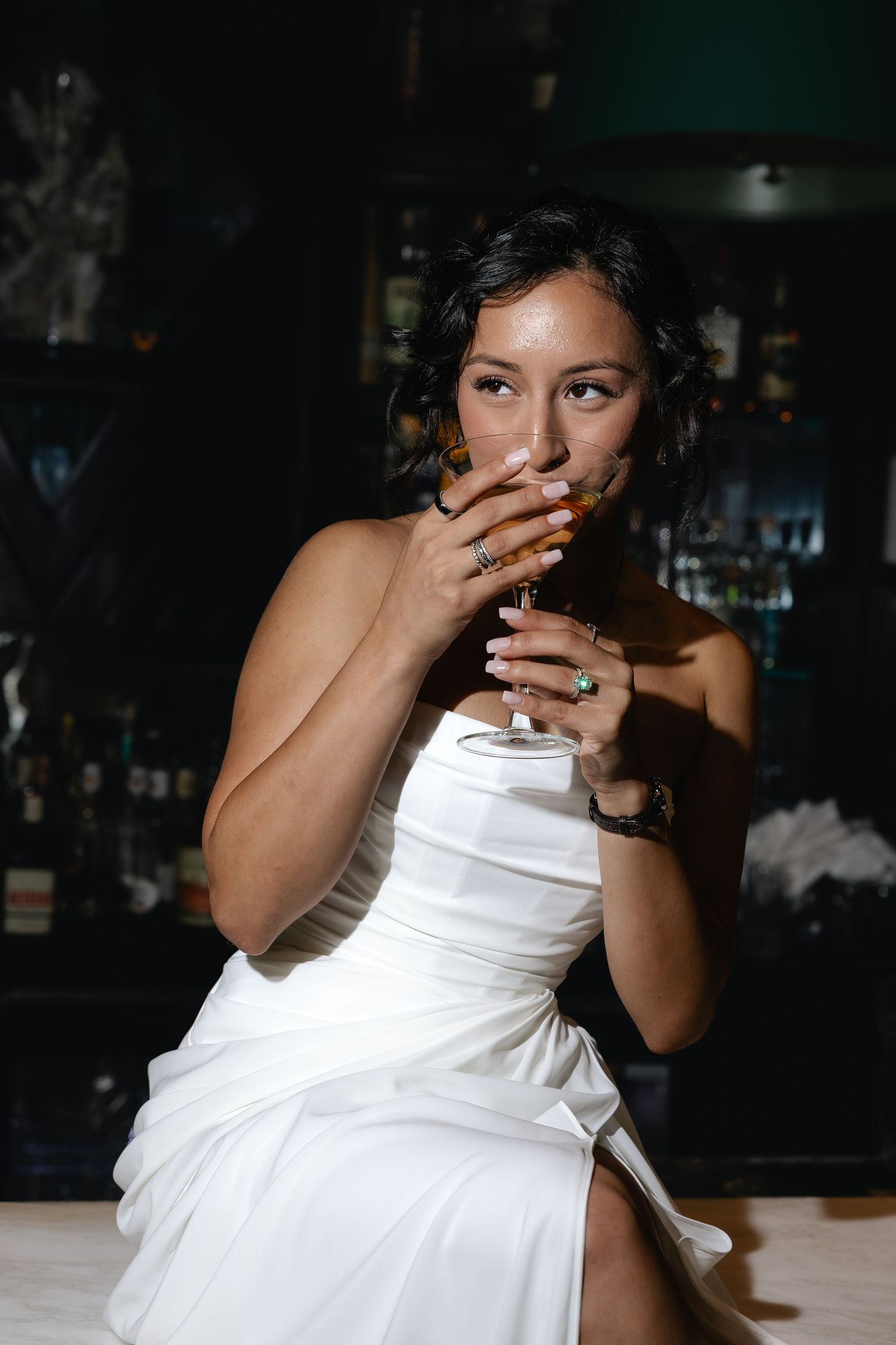 Woman in white strapless dress holding cocktail glass at dark bar, covering mouth with ringed hand.
