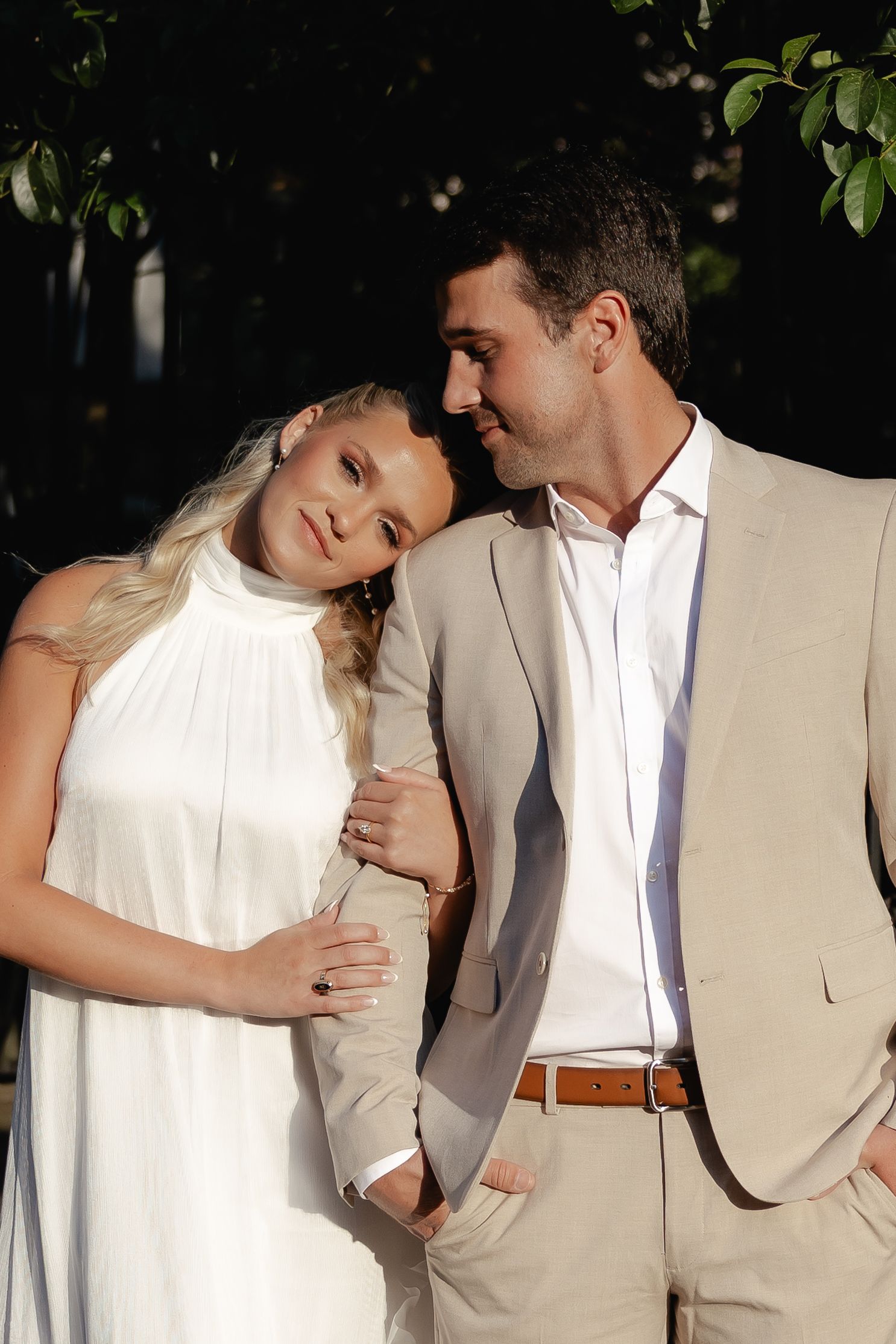 Bride in white halter dress leans on groom in tan suit while standing in dappled sunlight outdoors.