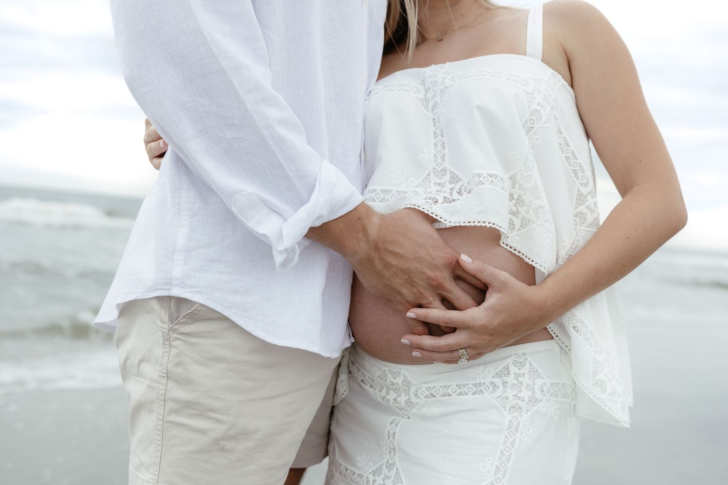 Pregnant couple in white clothing embracing on a beach, hands on baby bump.