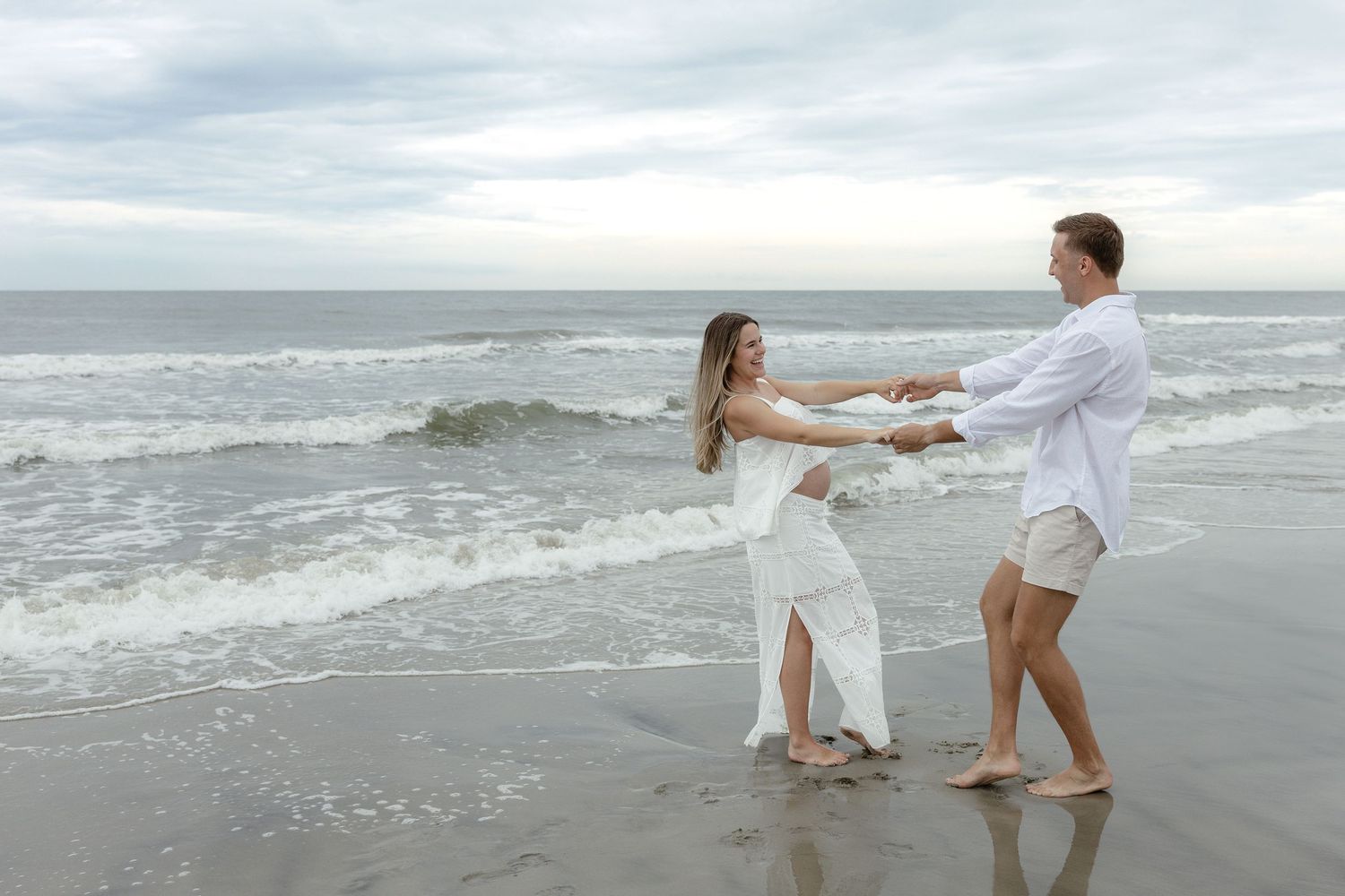 Couple in white outfits dancing on a beach with ocean waves in the background.
