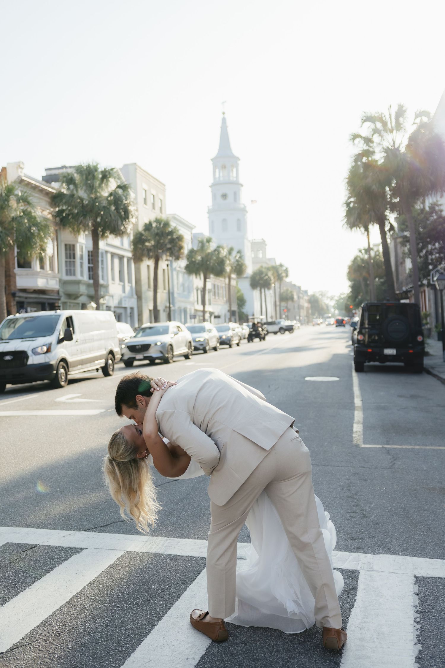 Groom dipping bride for a kiss on a sunny Charleston street with palm trees and church steeple.