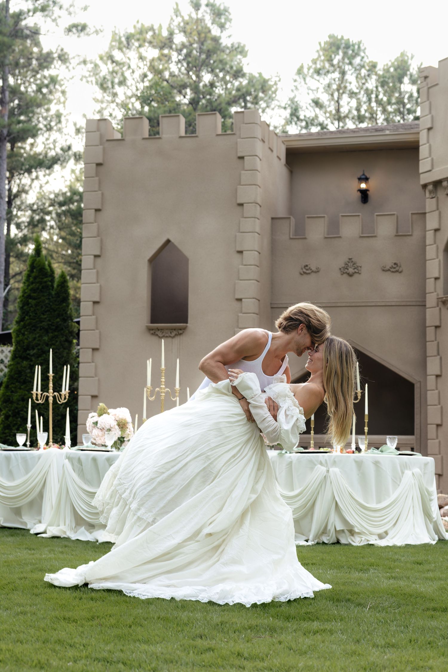 Couple sharing a romantic dip kiss in wedding attire outside a castle-style venue with elegant reception table.