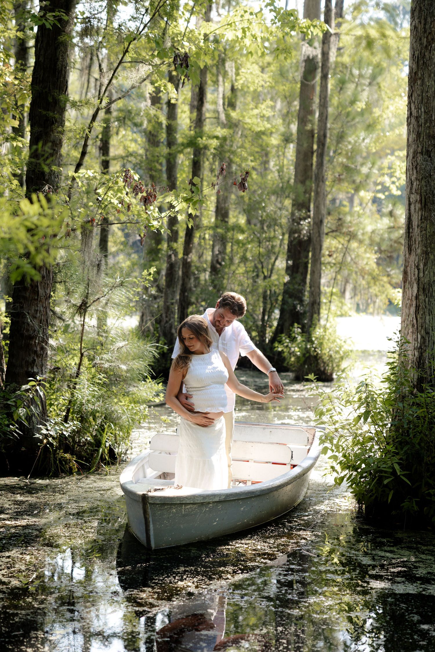 Romantic couple in white embracing in a rowboat surrounded by lush green trees in a tranquil swamp.