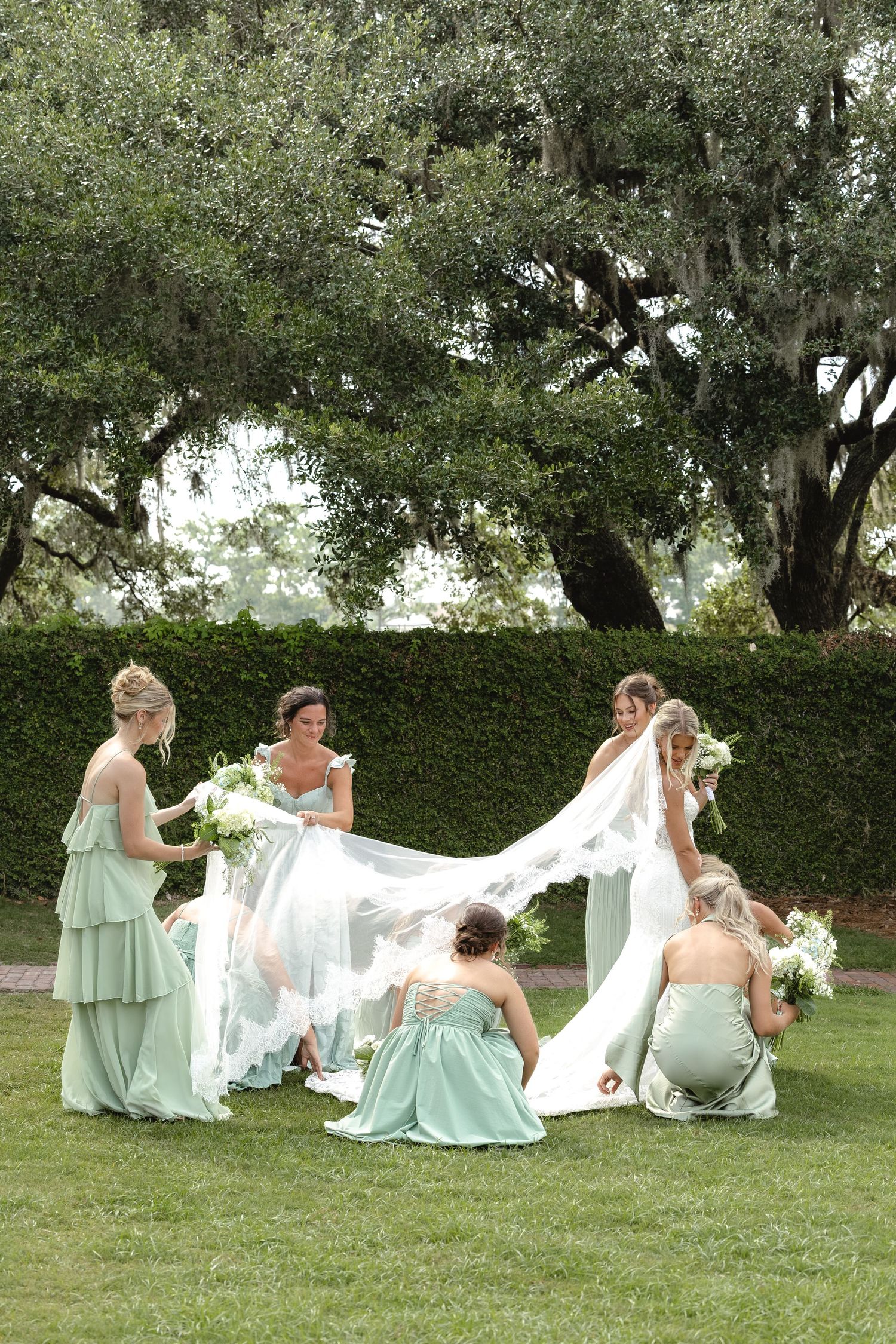 Bride surrounded by bridesmaids in mint green gowns arranging her white veil on a lush green lawn.