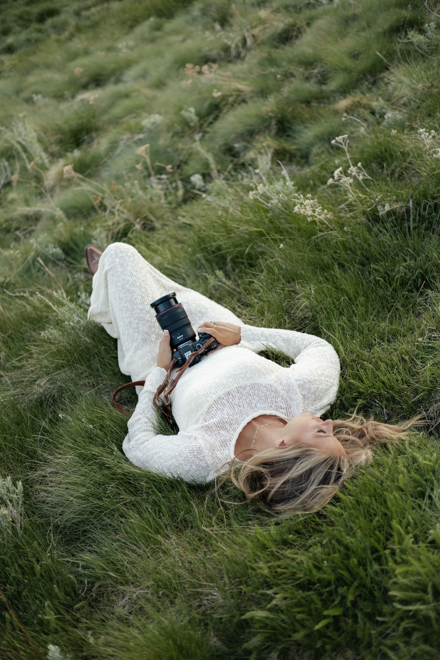 Woman in white lace outfit lying on lush green hillside grass holding a DSLR camera.