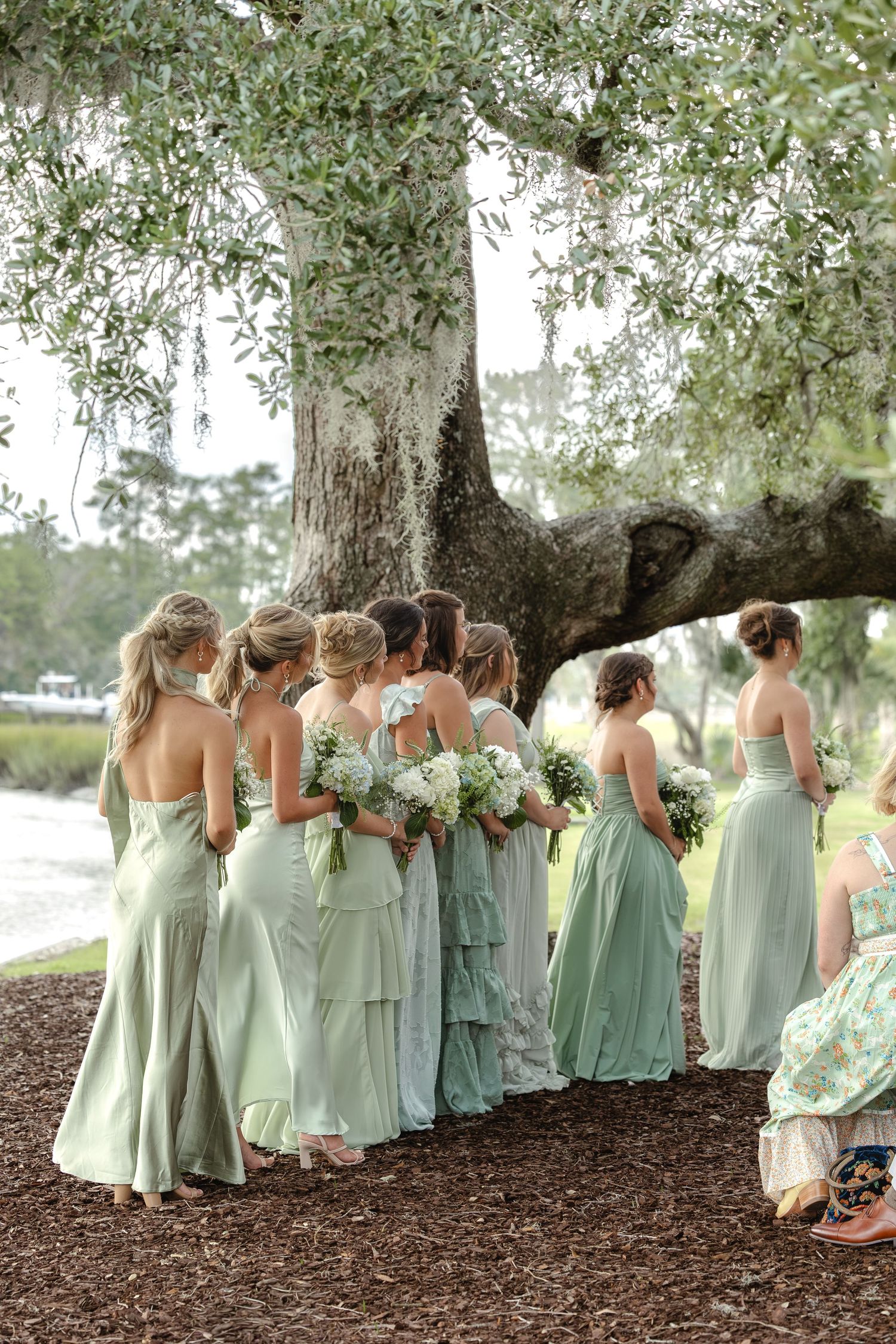Bridesmaids in sage green gowns holding white bouquets stand under a large oak tree at an outdoor wedding.