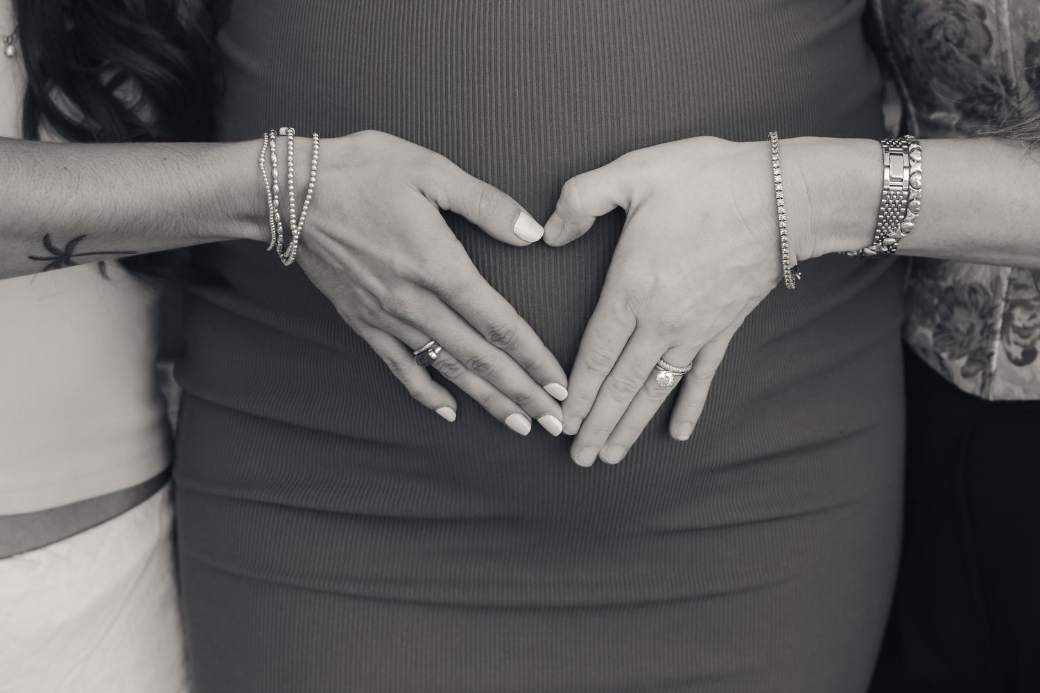 Two people form a heart shape with their hands over a pregnant belly in black and white.