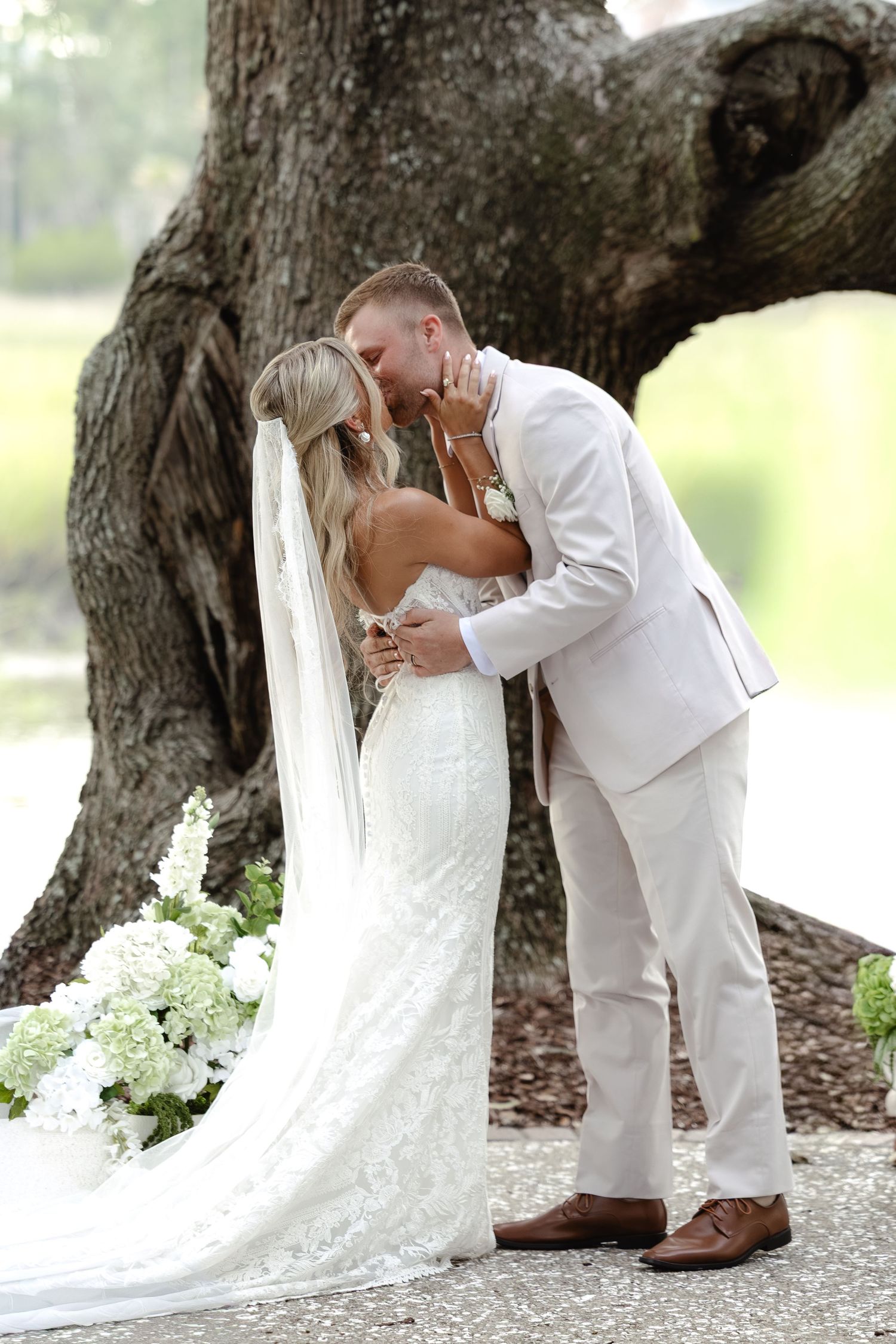 Bride and groom share a romantic first kiss under a large oak tree, bride wearing lace gown with long veil.