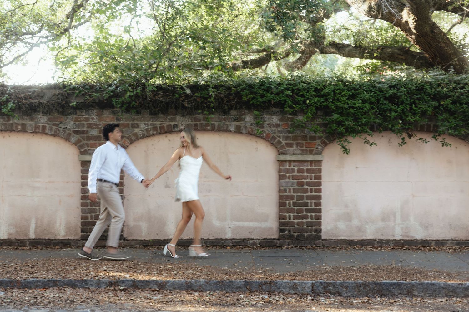 Couple holding hands and walking along a brick wall covered in ivy and vines on a sunny day.