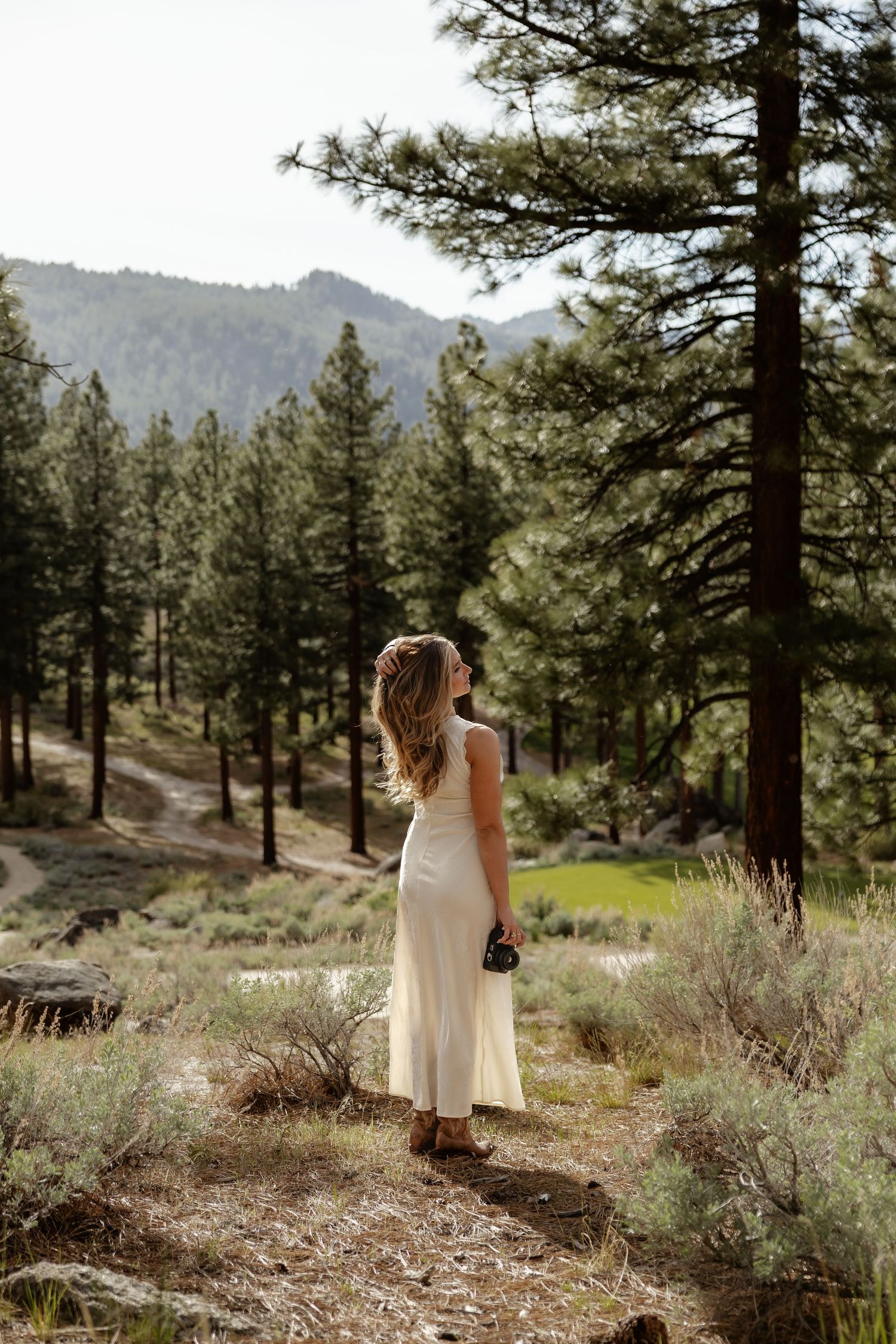 Blonde woman in white dress holding a camera stands on a forest trail surrounded by pine trees and mountains.