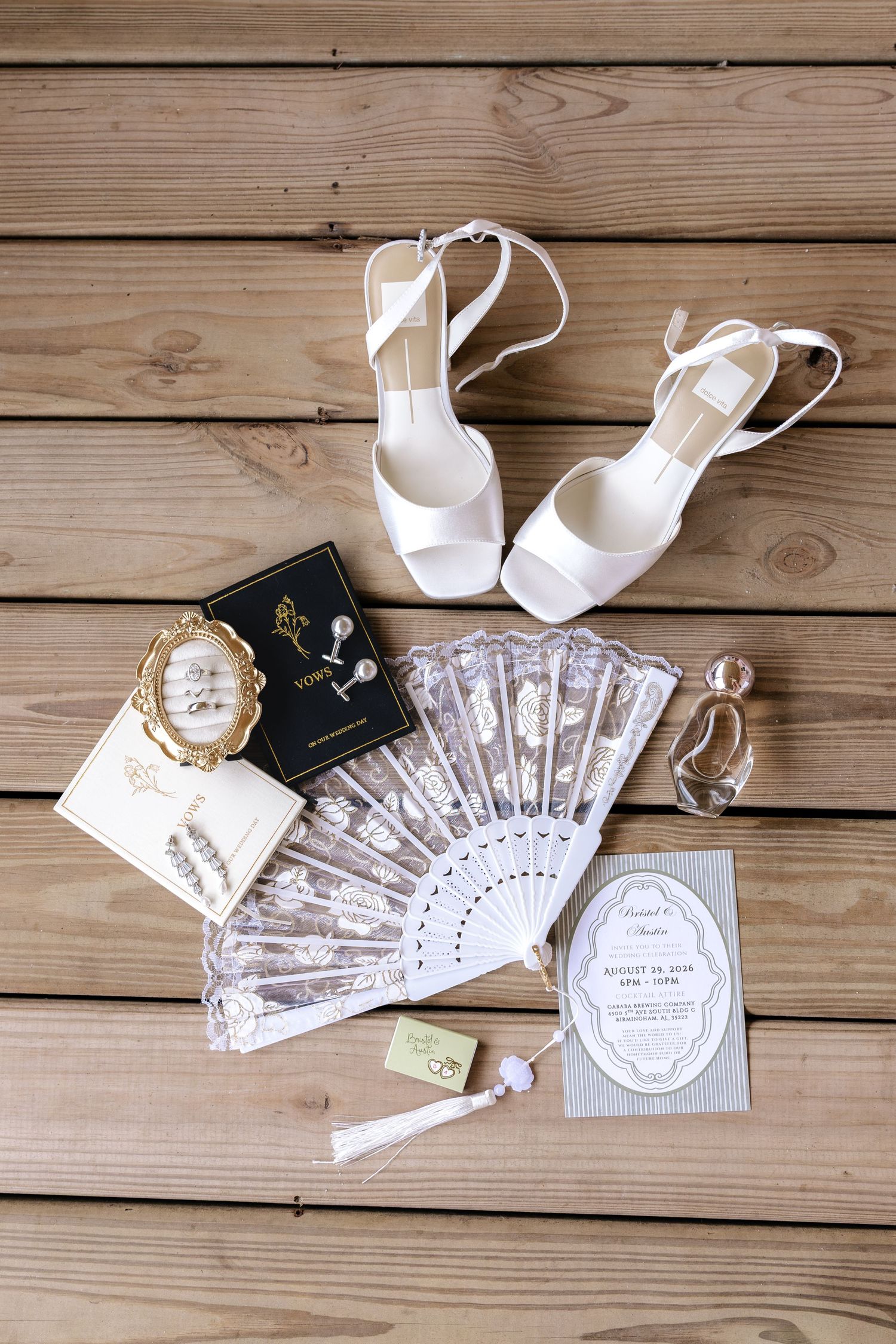 White bridal heels, lace fan, perfume, invitations, and accessories arranged on rustic wooden planks for a wedding flat lay.