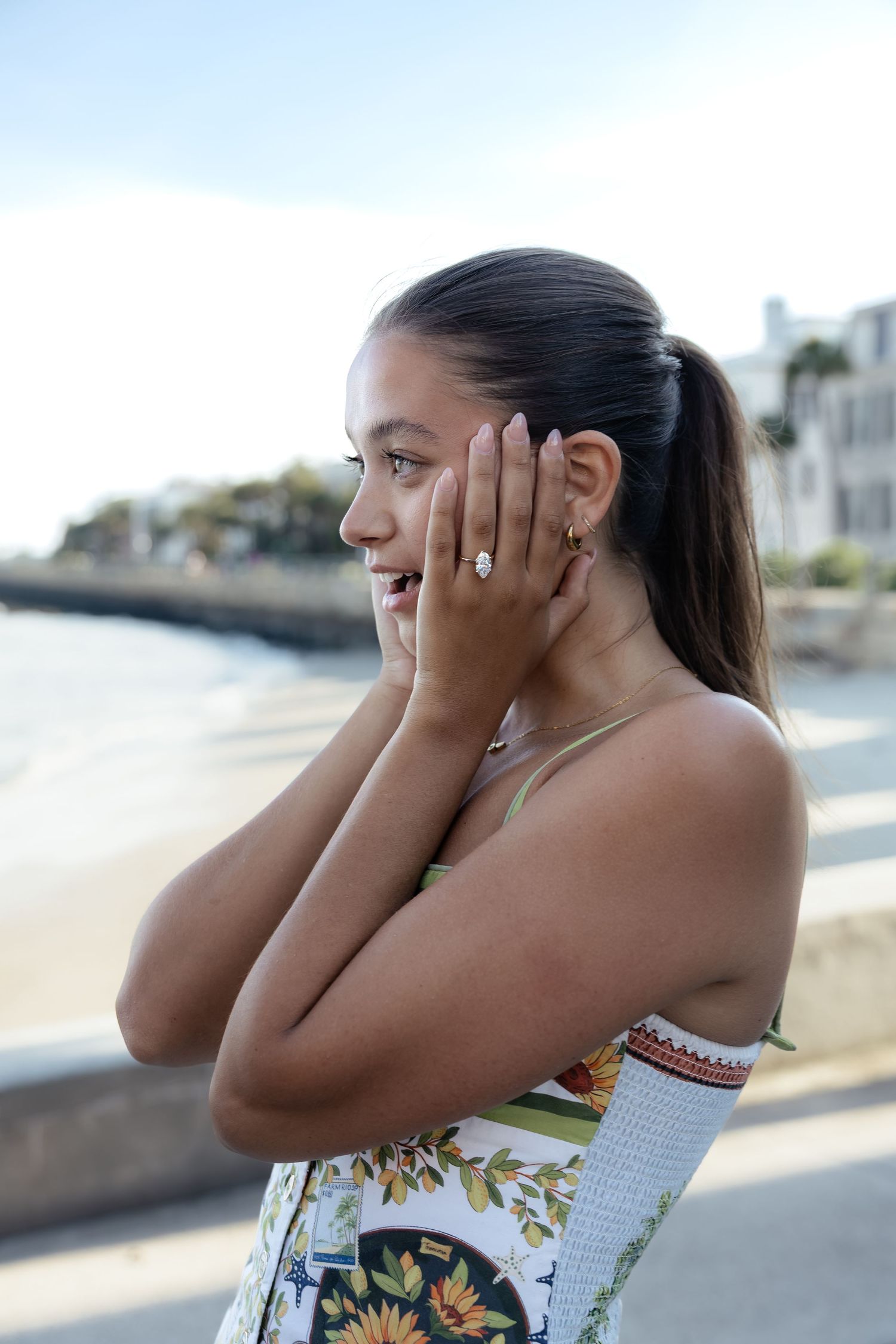 Young woman in floral dress admiring her engagement ring on a sunny waterfront promenade.