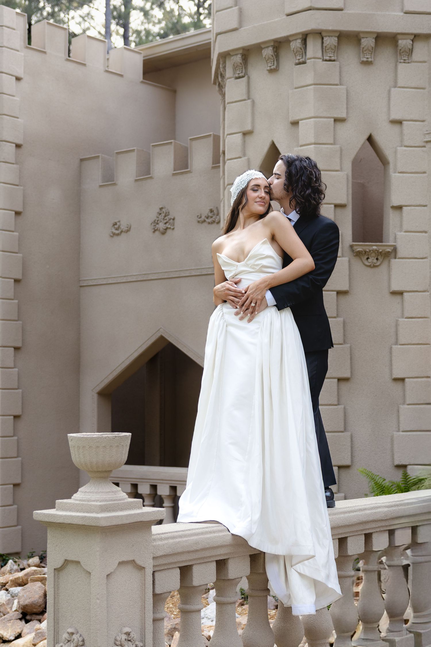 Bride and groom embrace romantically on a castle balcony, with the bride in a white gown and groom in a dark suit.