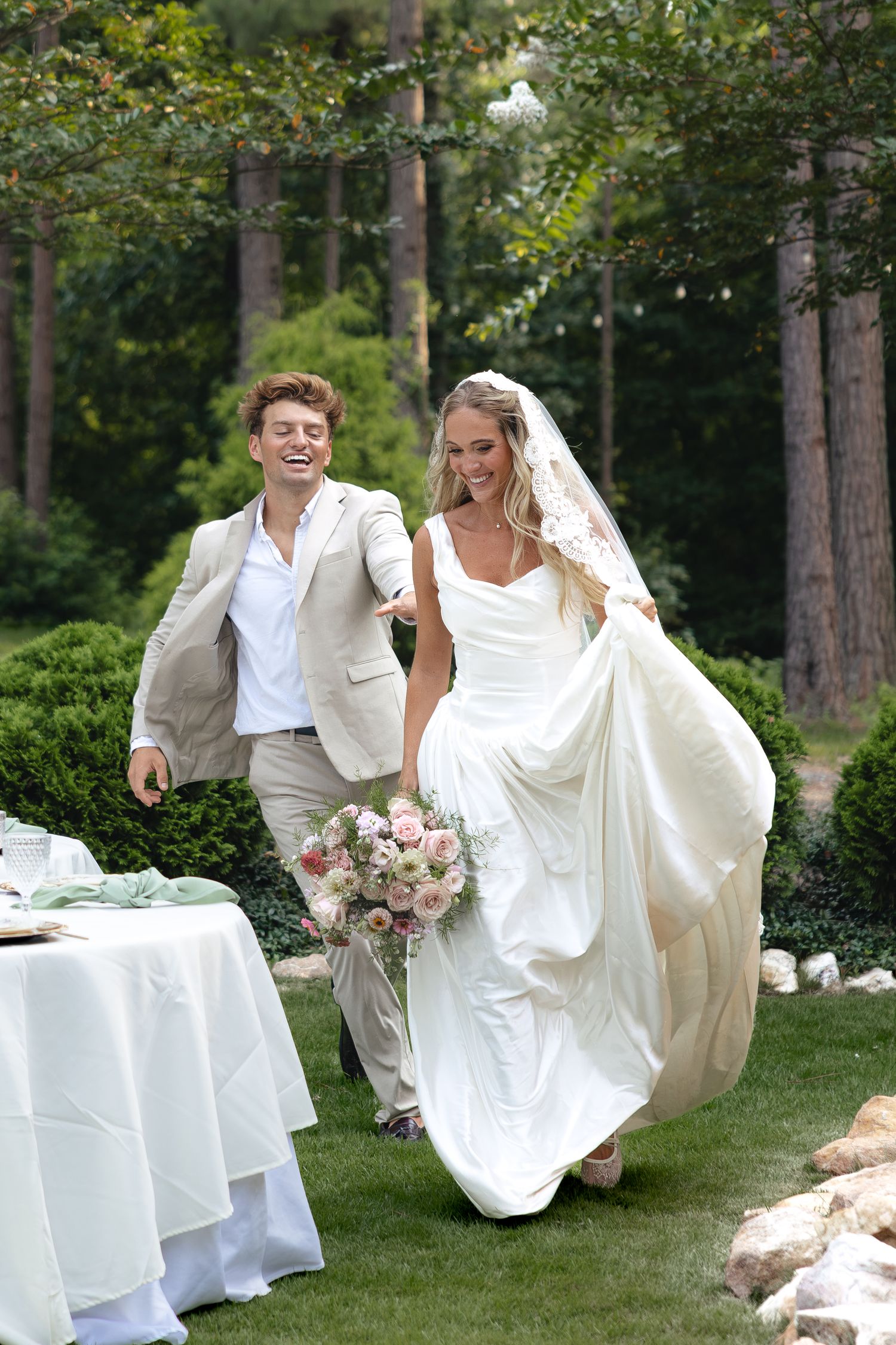 Joyful bride in white gown holding pink bouquet walks with laughing groom in beige suit at outdoor wedding.