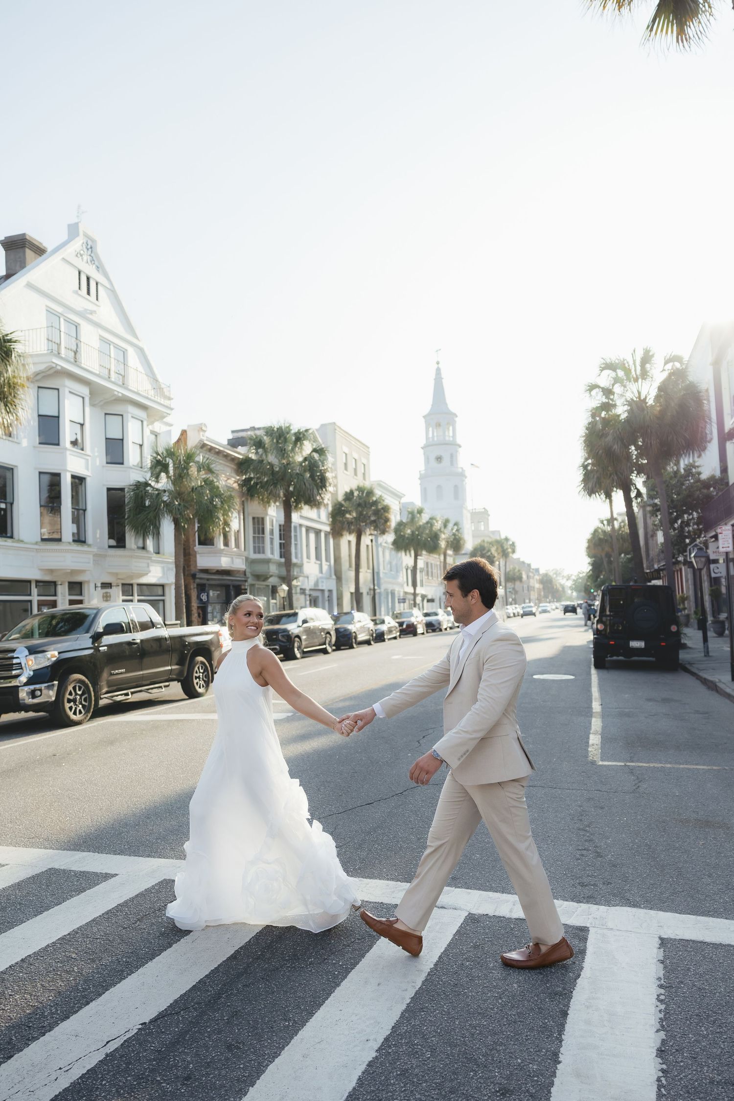 Bride and groom hold hands crossing a sunny Charleston street with palm trees and church steeple backdrop.