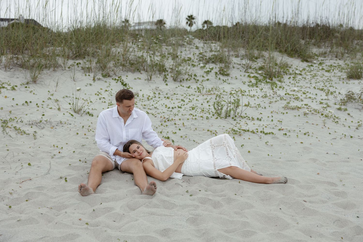 Couple relaxing on sandy beach dunes, woman lying with head on man's lap in matching white outfits.